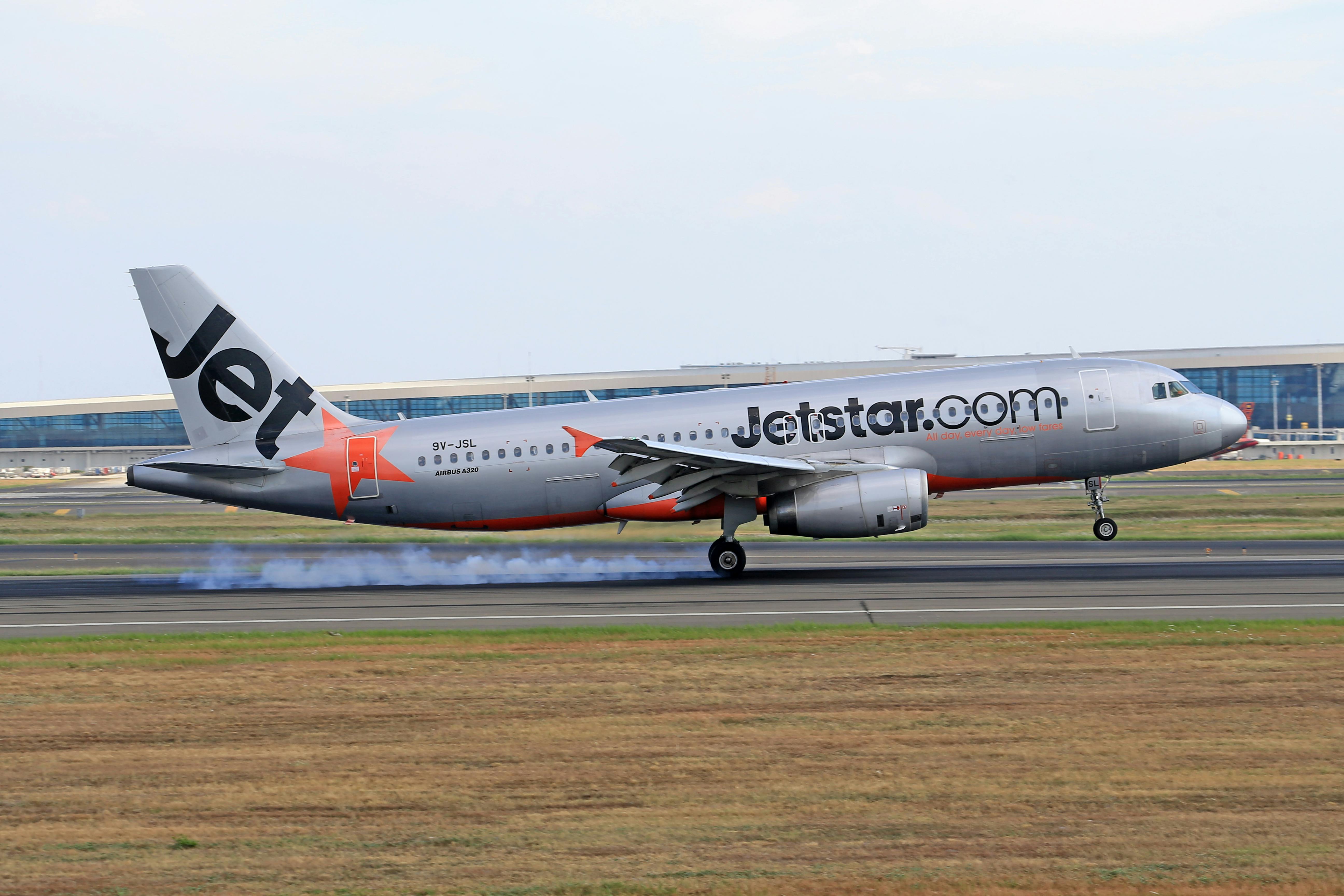 Jetstar Airbus A320 landing with visible tire smoke on runway near a terminal.