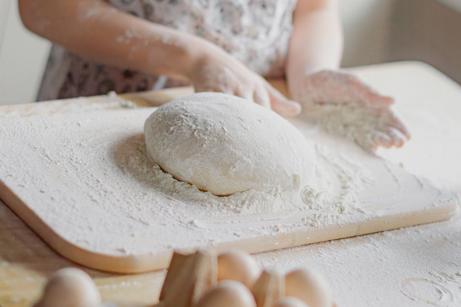 A woman preparing dough on a wooden table in a bright kitchen, hands covered in flour.