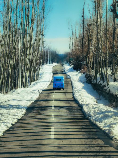 Free A vibrant blue van driving down a snow-covered rural road in Anantnag during winter, framed by bare trees. Stock Photo