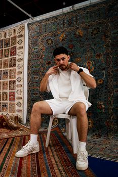A stylish man in white attire sitting against ornate Persian rugs.