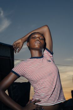 A young woman in a striped shirt poses confidently against a sunset backdrop.