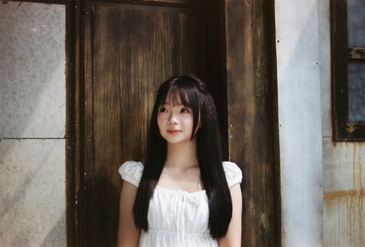 Young woman in a white dress stands by a rustic wooden door, creating a serene and classic portrait.