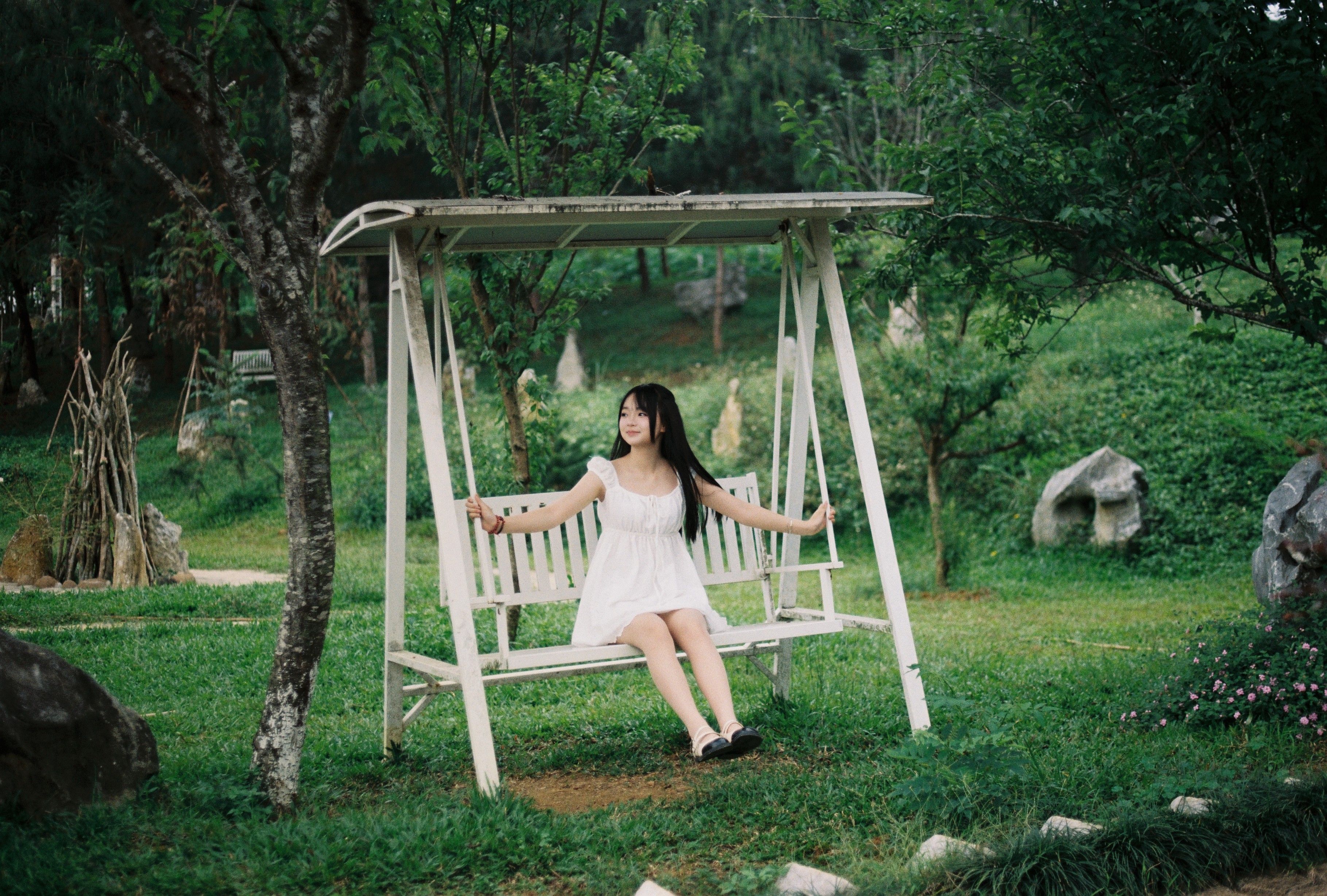 A young woman in a white dress relaxing on a swing in a lush, green garden.
