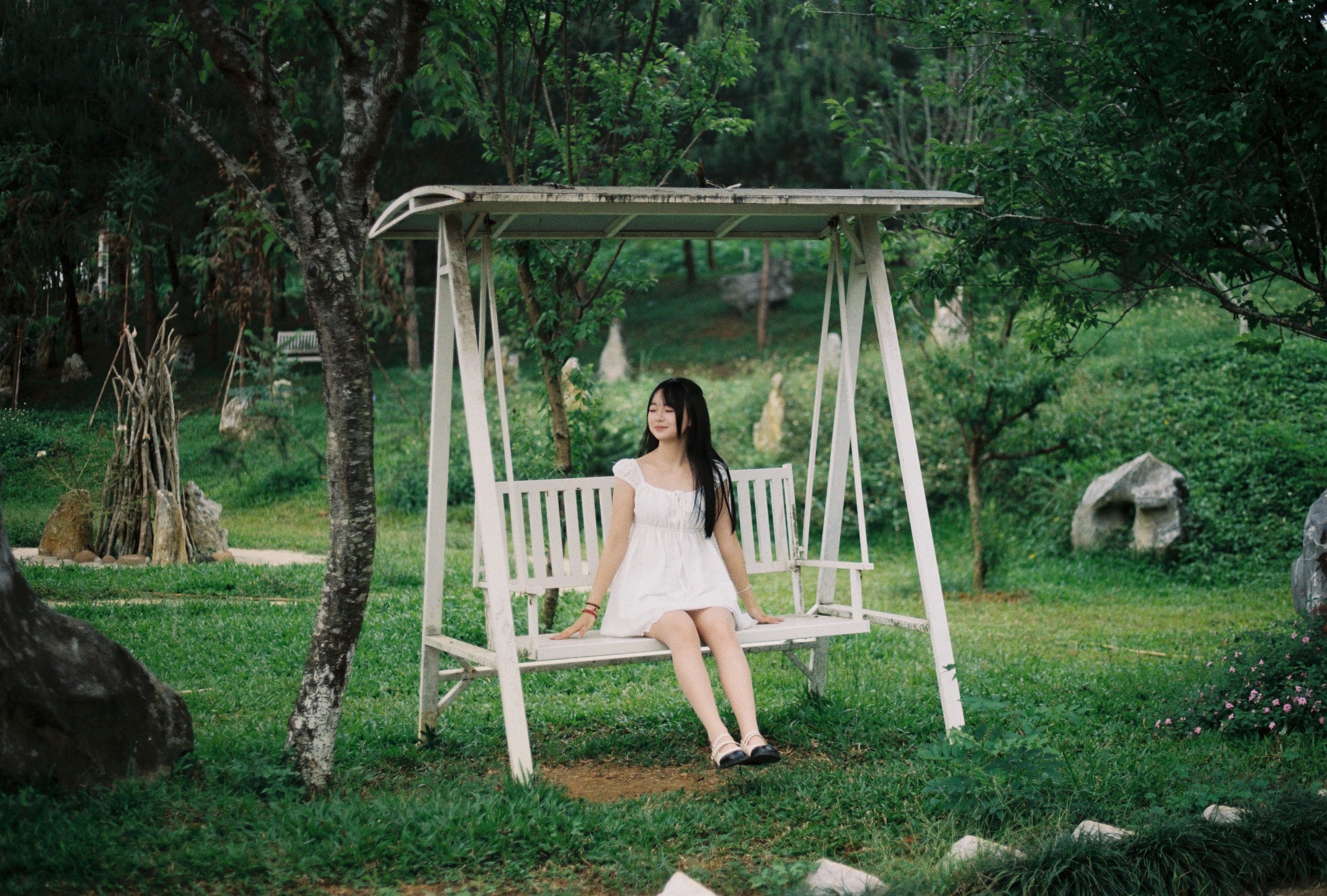 A young woman in a white dress sits peacefully on a garden swing surrounded by lush greenery.