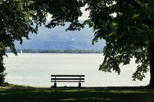 Tranquil scene of an empty bench overlooking Bodensee with a distant sailboat under leafy trees.