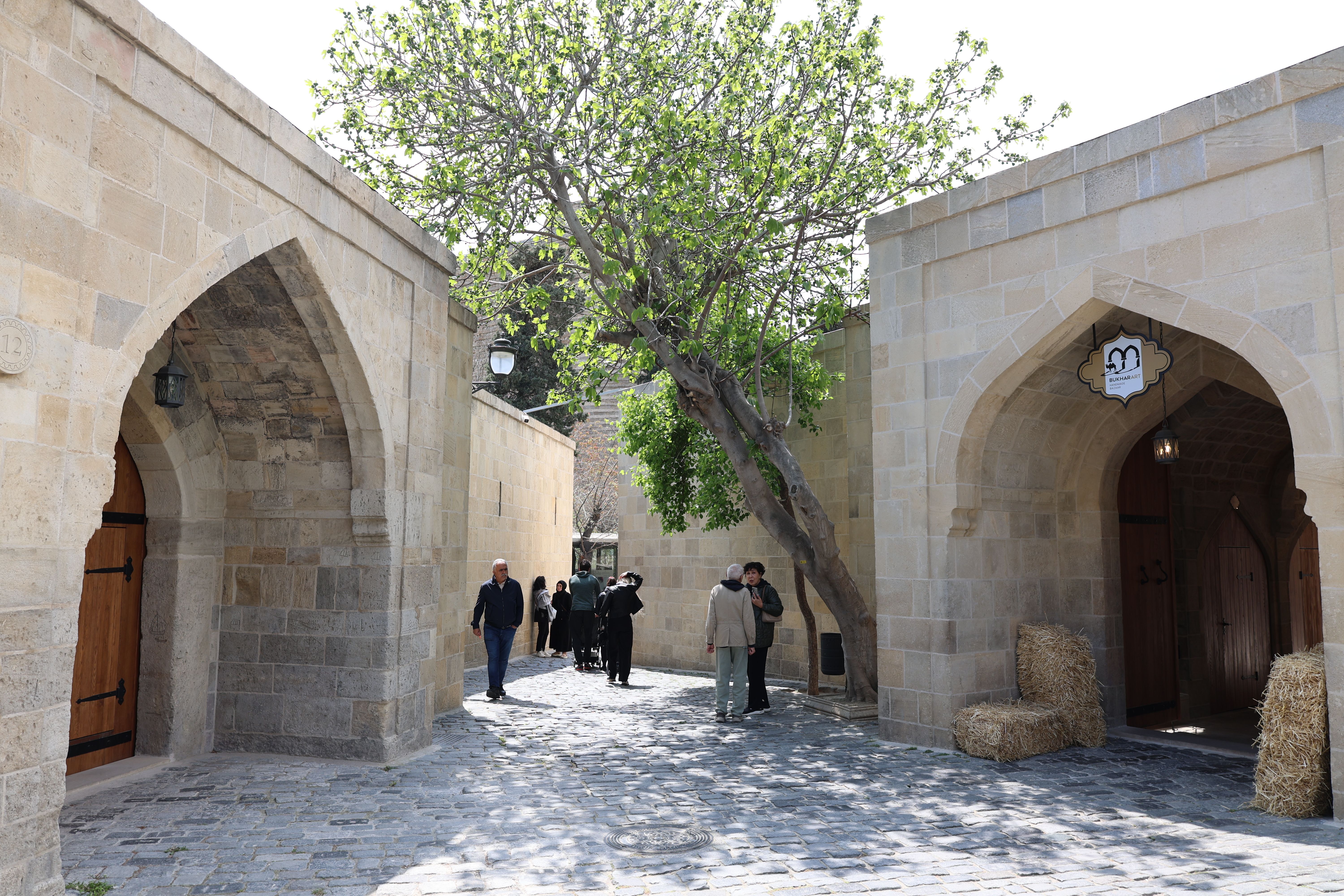 Free A serene courtyard with stone buildings, archways, and a large tree, featuring a few people walking. Stock Photo