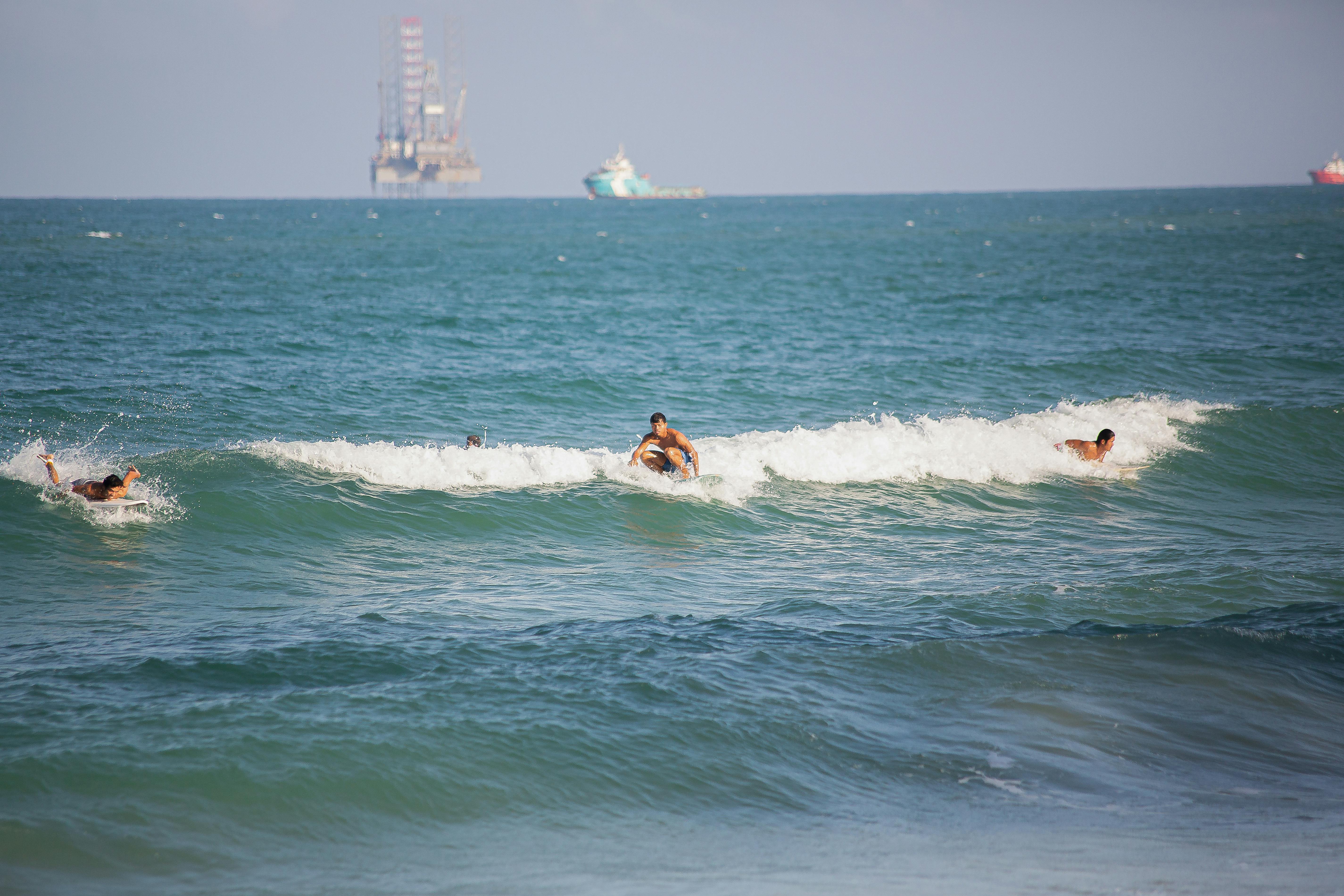 Surfers enjoy waves with an offshore drilling rig in the background, under a clear sky.