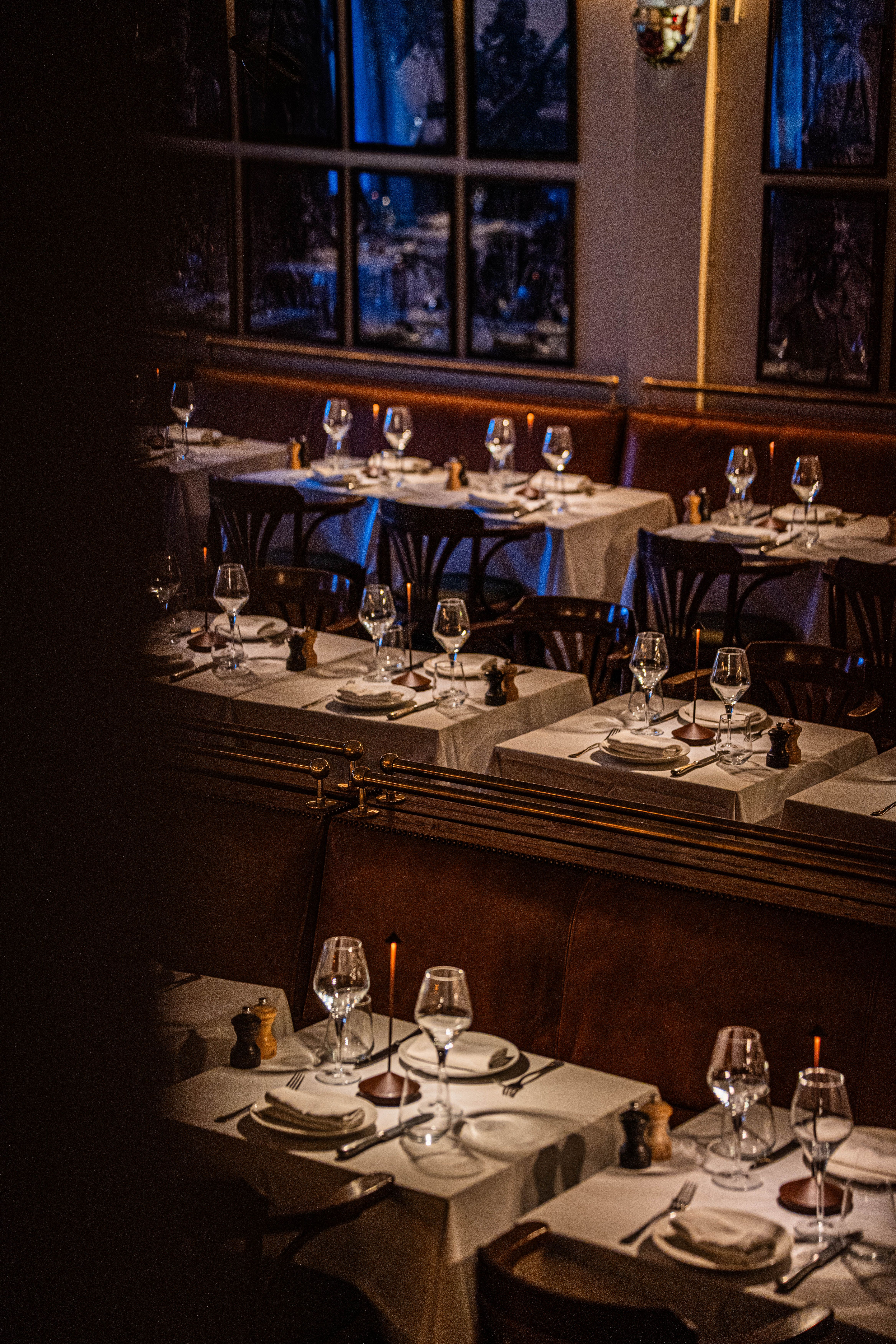 Salle de restaurant en soirée : tables nappées de blanc, verres à pied alignés et chaises en bois, lumière tamisée