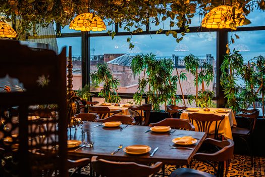 Chic restaurant interior featuring tables set for dining amid lush greenery, with cityscape visible through large windows.
