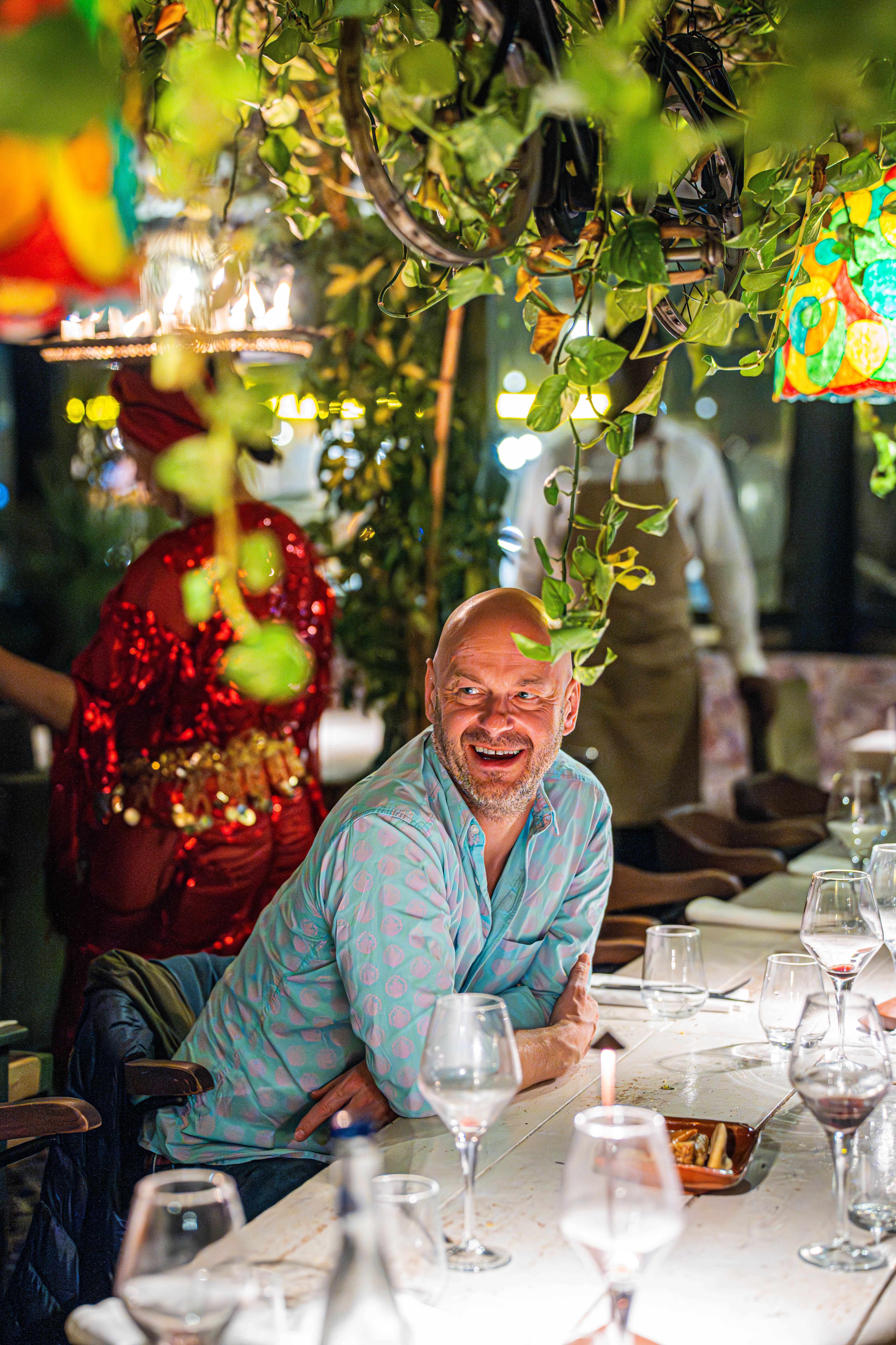 A cheerful man enjoying a lively dinner with friends in a beautifully decorated restaurant with hanging plants.