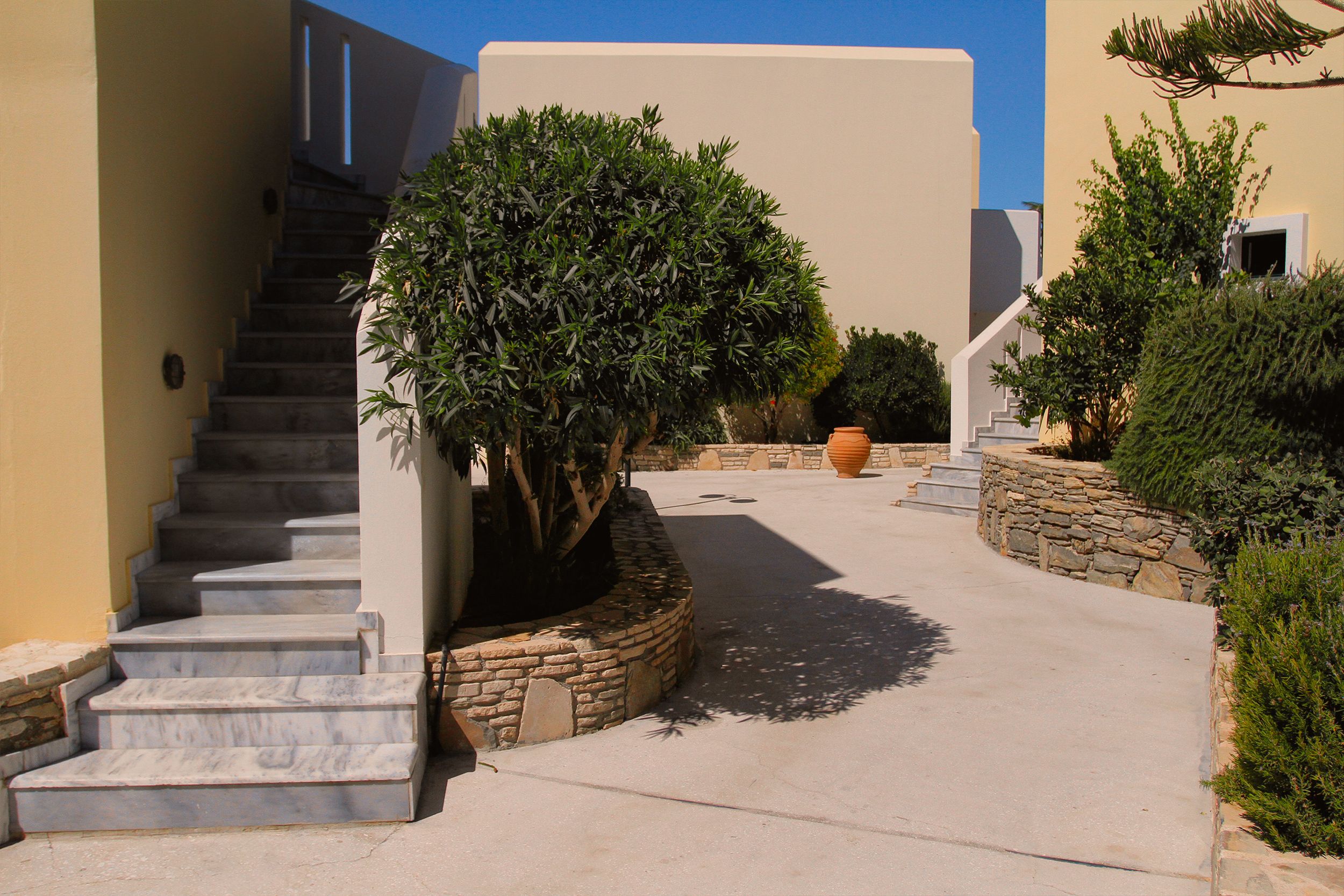 A serene Mediterranean courtyard featuring marble stairs and lush greenery under a clear blue sky.
