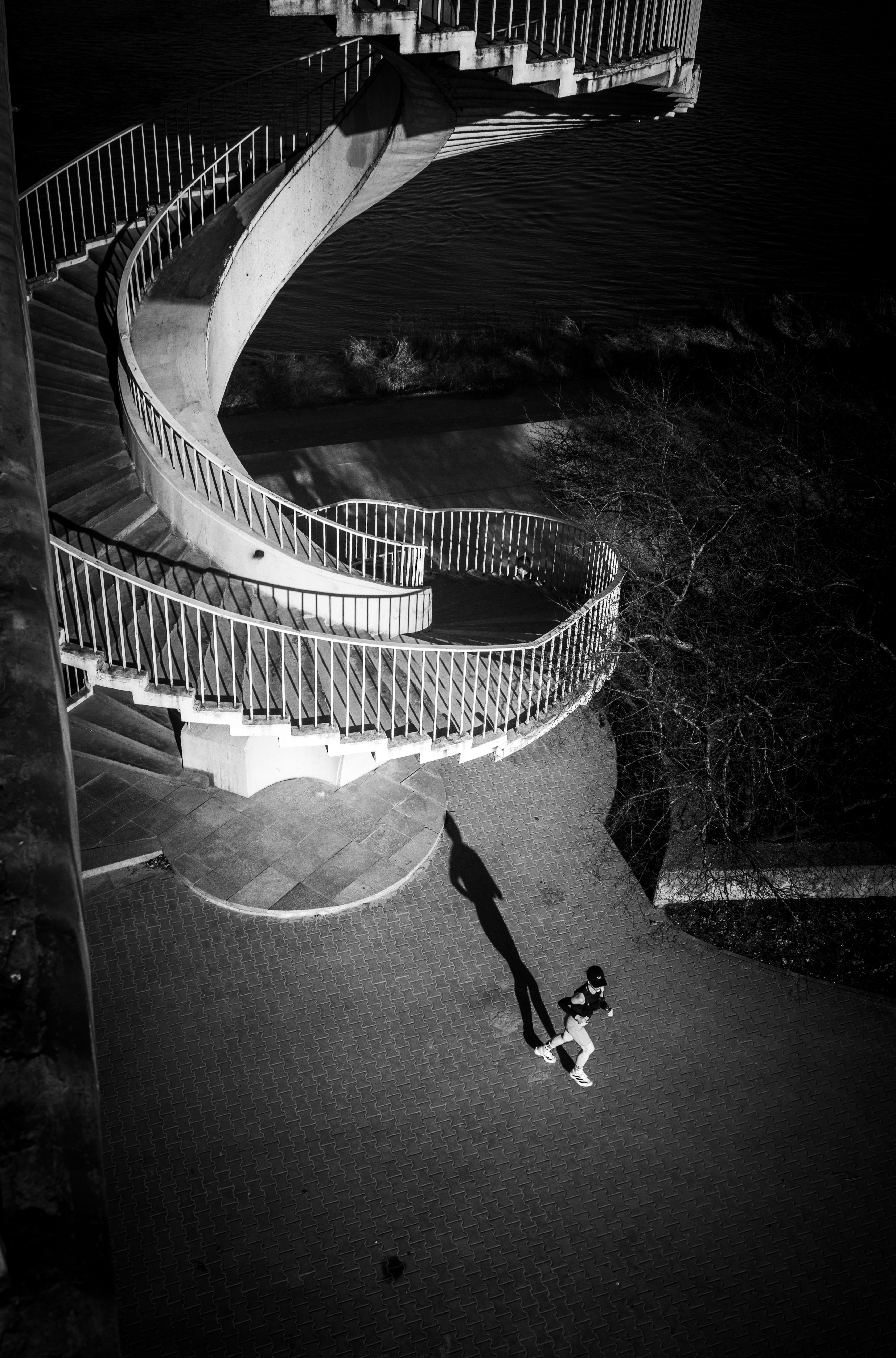 A dramatic black and white aerial shot of a winding staircase casting long shadows with a lone walker below.