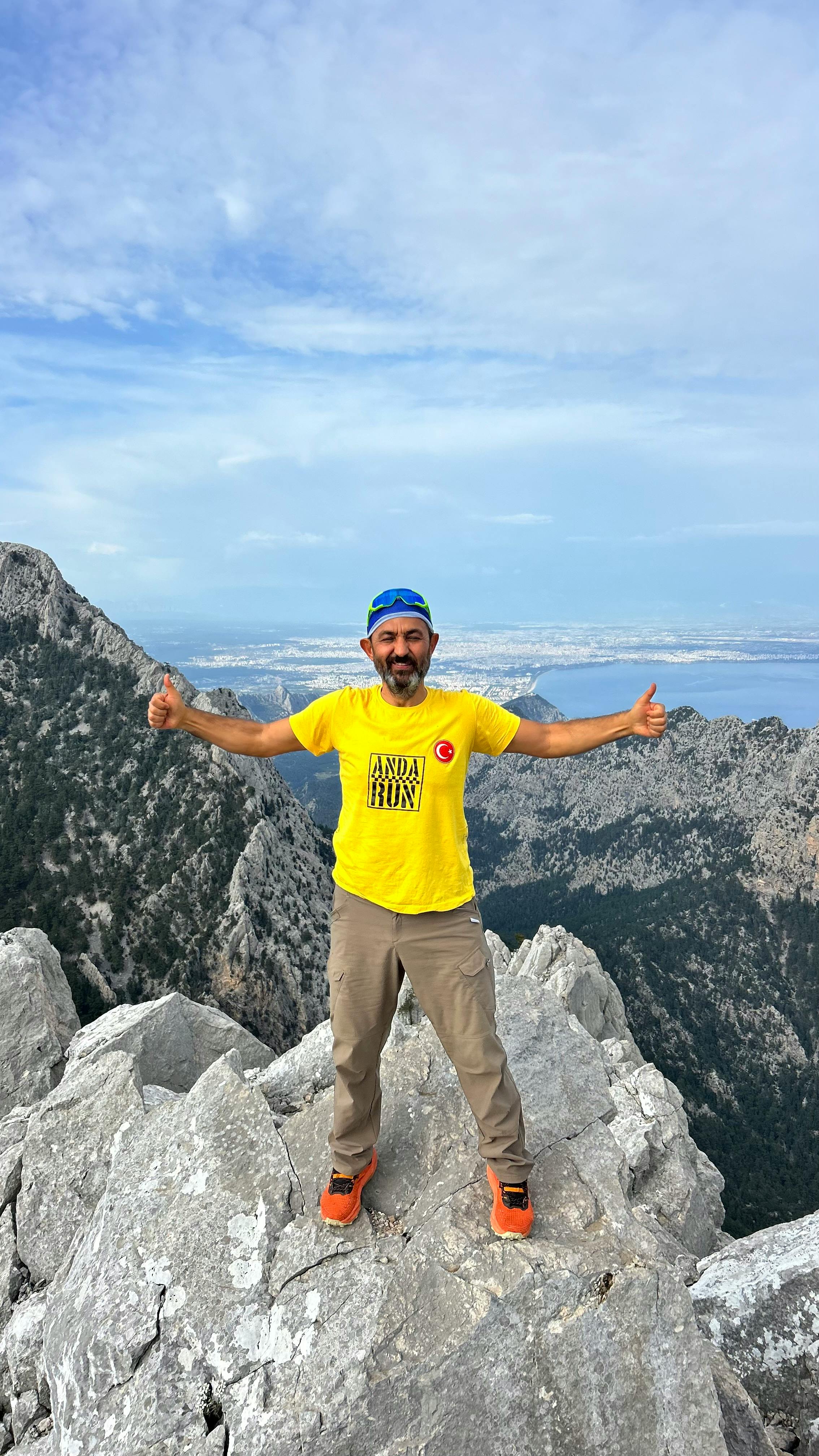 A man in a yellow shirt celebrates on a rocky mountain summit, with expansive views behind.