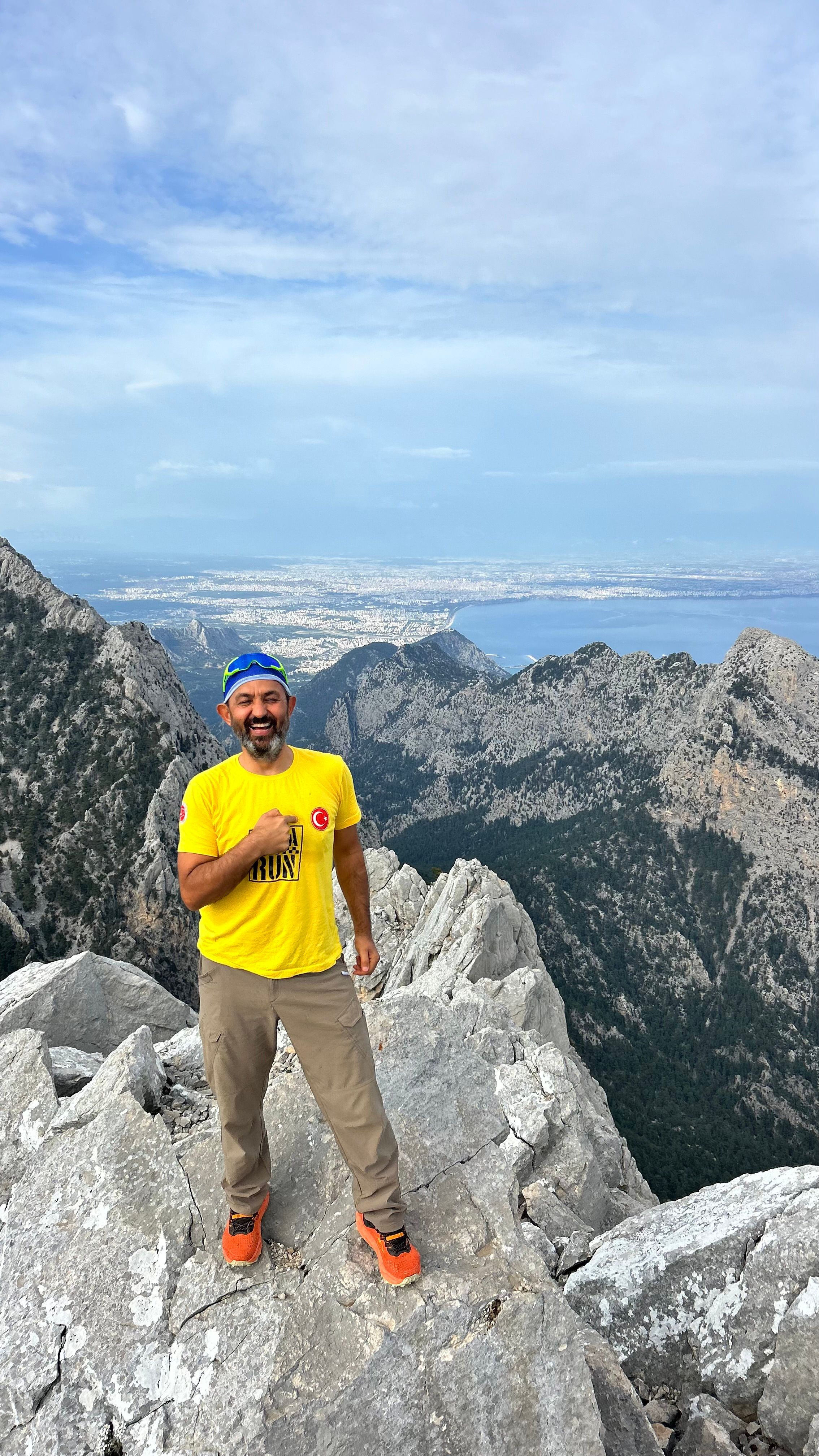 A hiker in a bright yellow t-shirt smiles atop a rocky mountain peak under a clear sky.