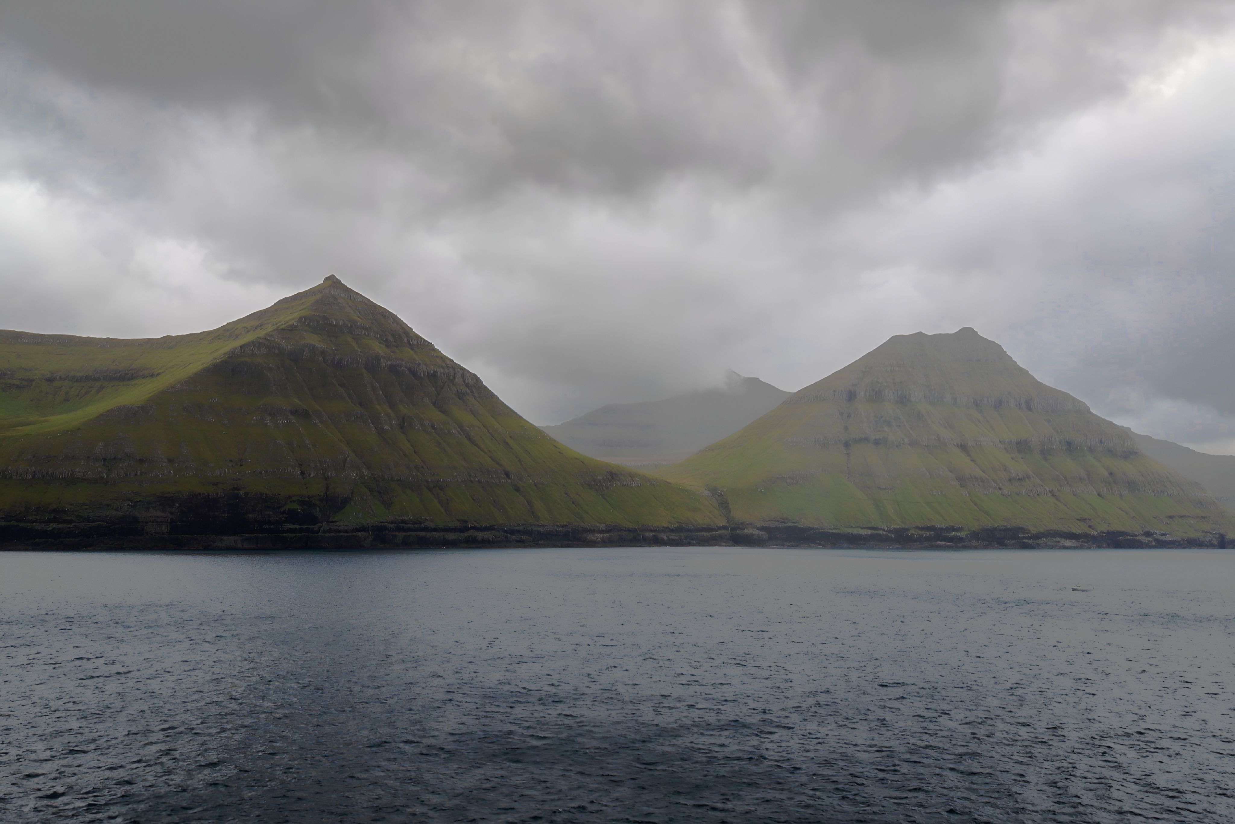 Misty hills under a cloudy sky in the Faroe Islands, reflecting a dramatic landscape over the water.