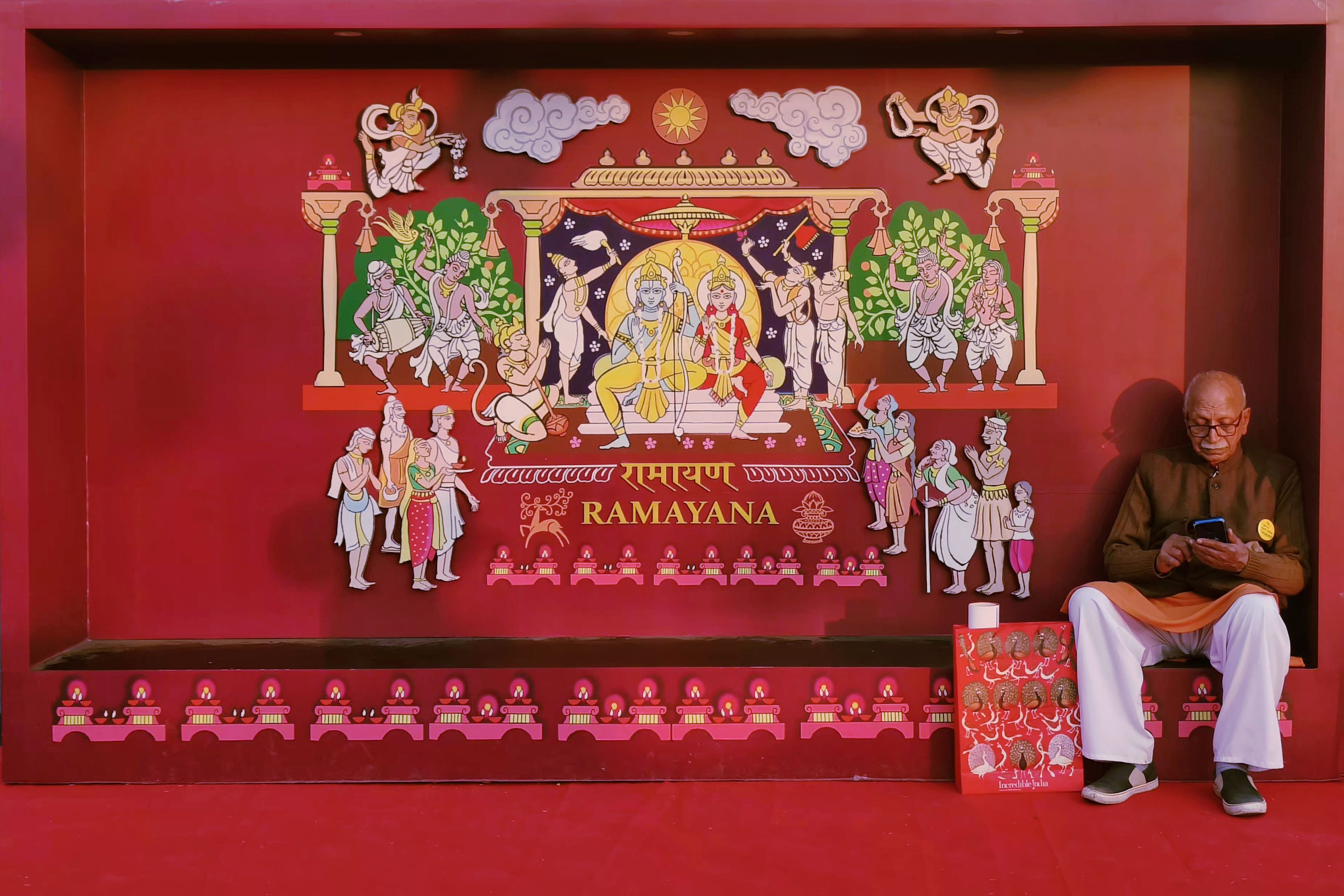Elderly man seated by vibrant Ramayana mural engrossed in phone, cultural depiction.