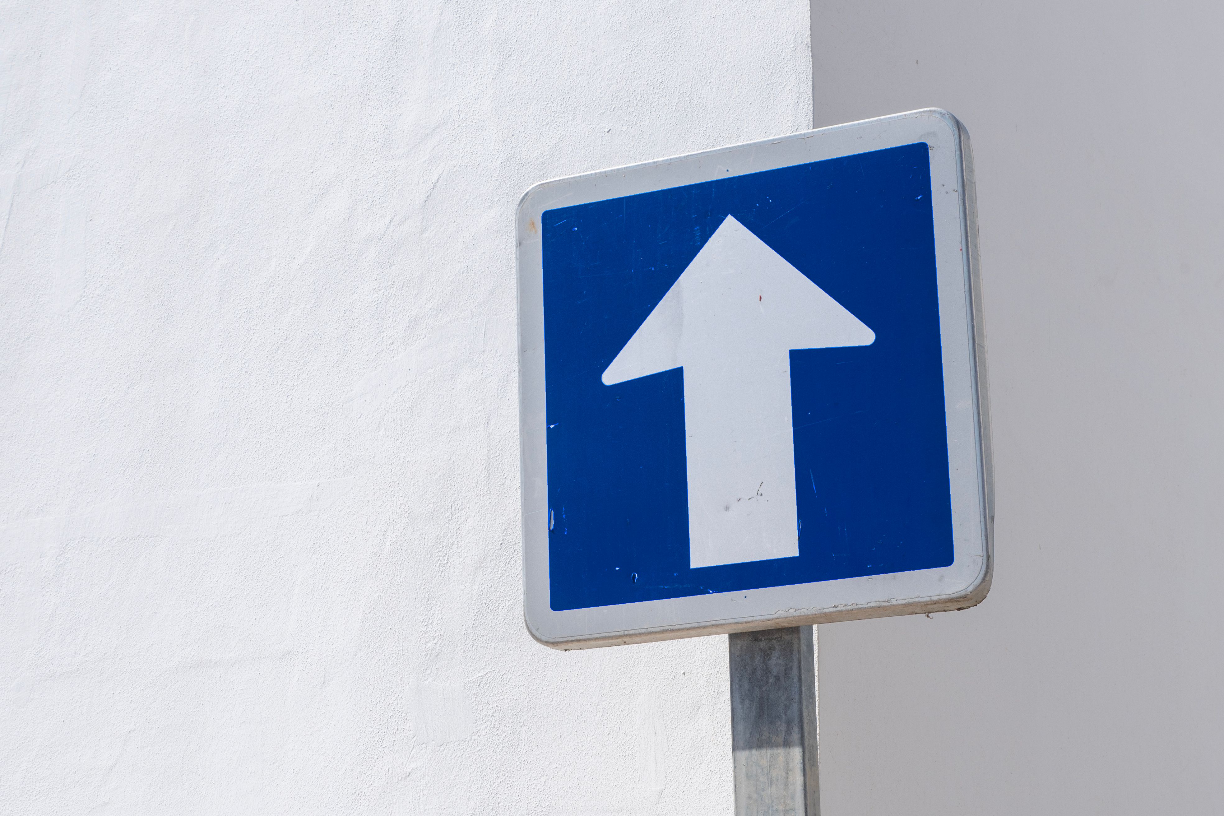 A simple blue directional traffic sign with white arrow over a white wall background, symbolizing direction or movement.