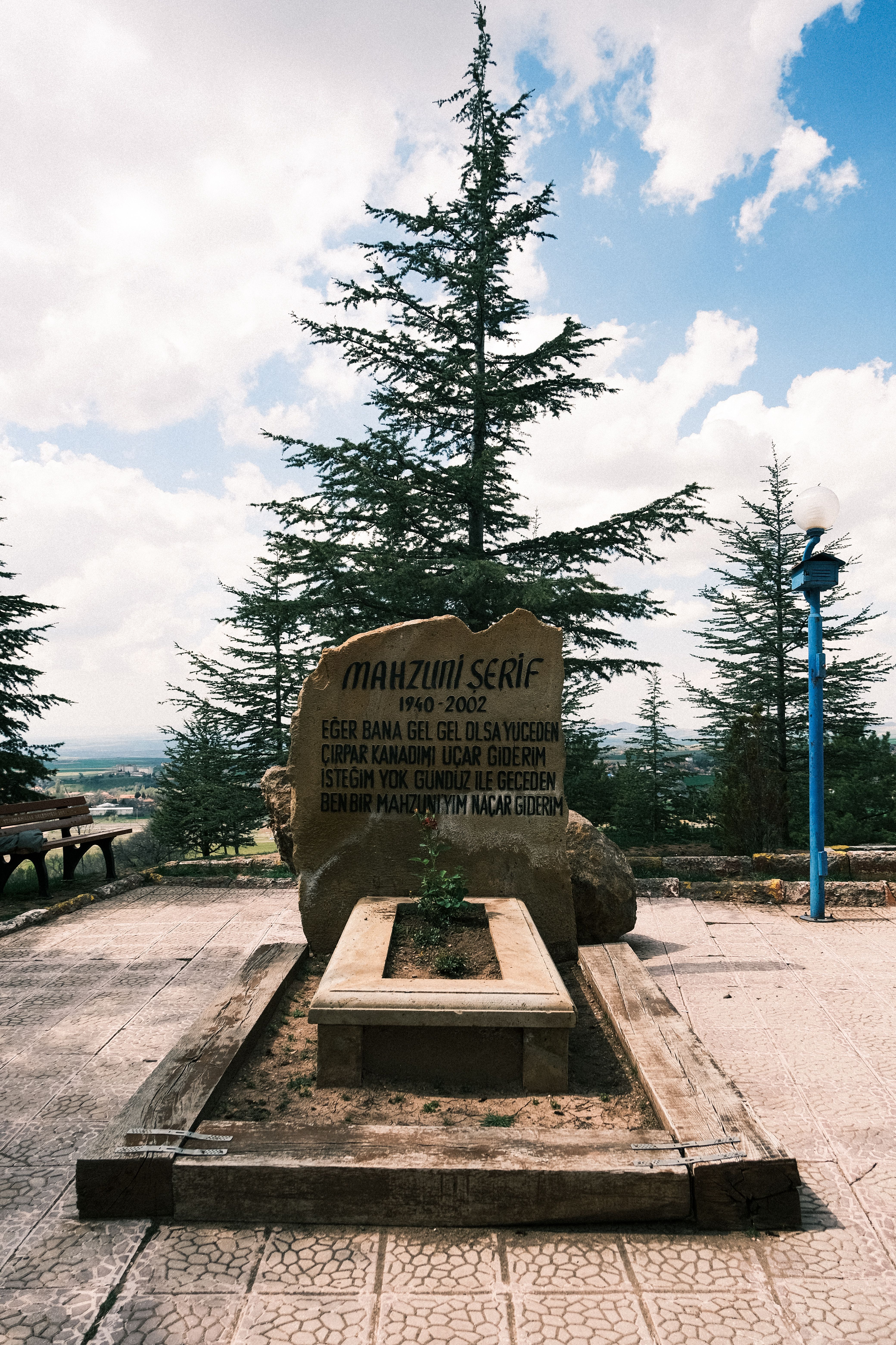 Memorial stone in a park setting under a sunny sky with evergreen trees.