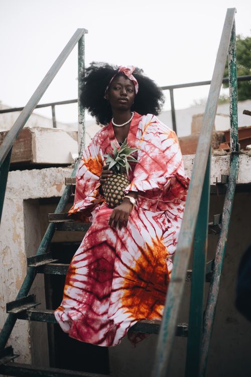 Free Stylish African woman wearing tie-dye clothes, seated on stairs with pineapple. Stock Photo