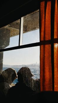 Silhouetted passengers looking out a window over a beautiful waterfront view through droplets-covered glass.