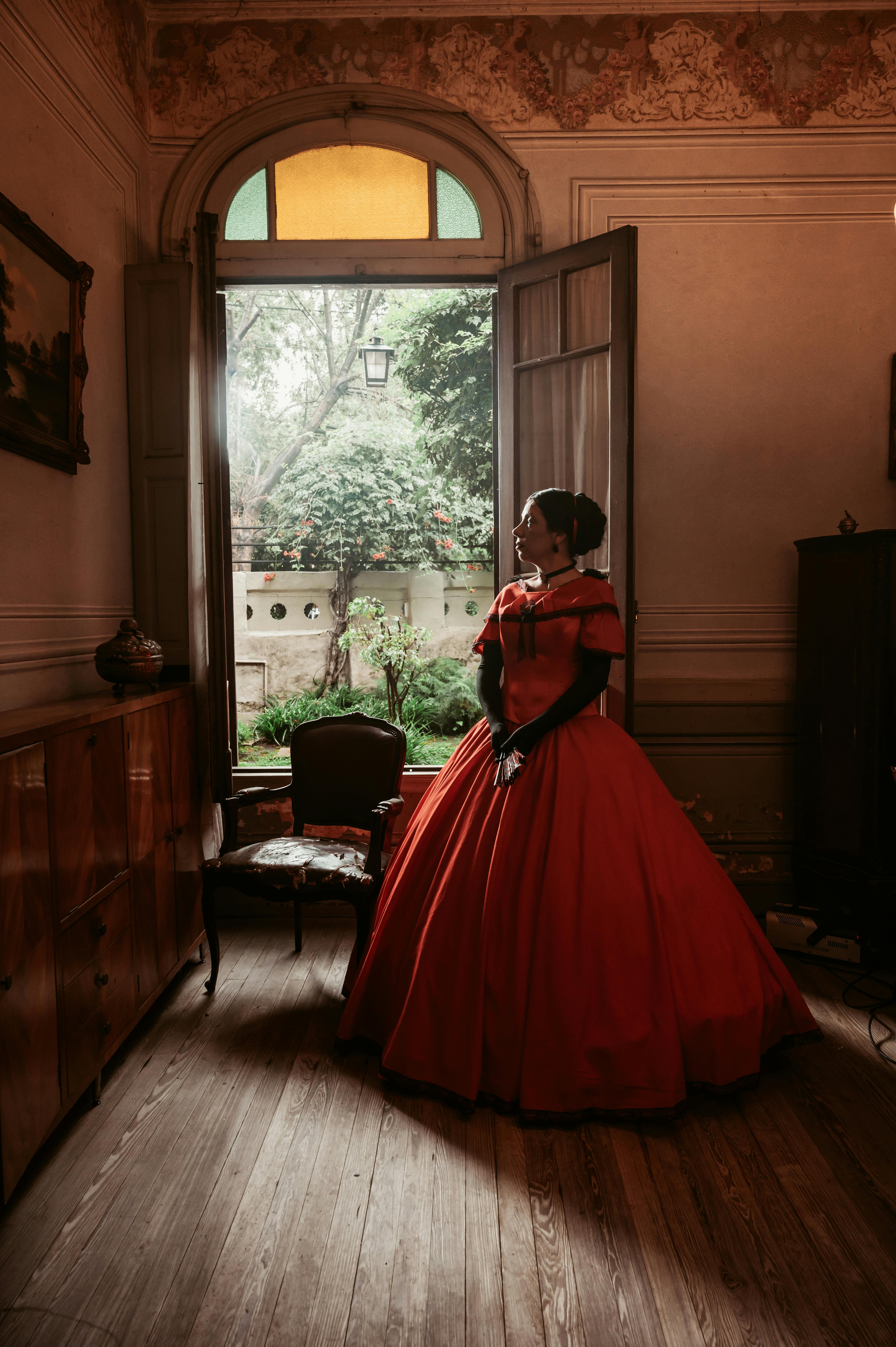 Elegant Victorian woman in red gown gazing out an open window in a vintage room.