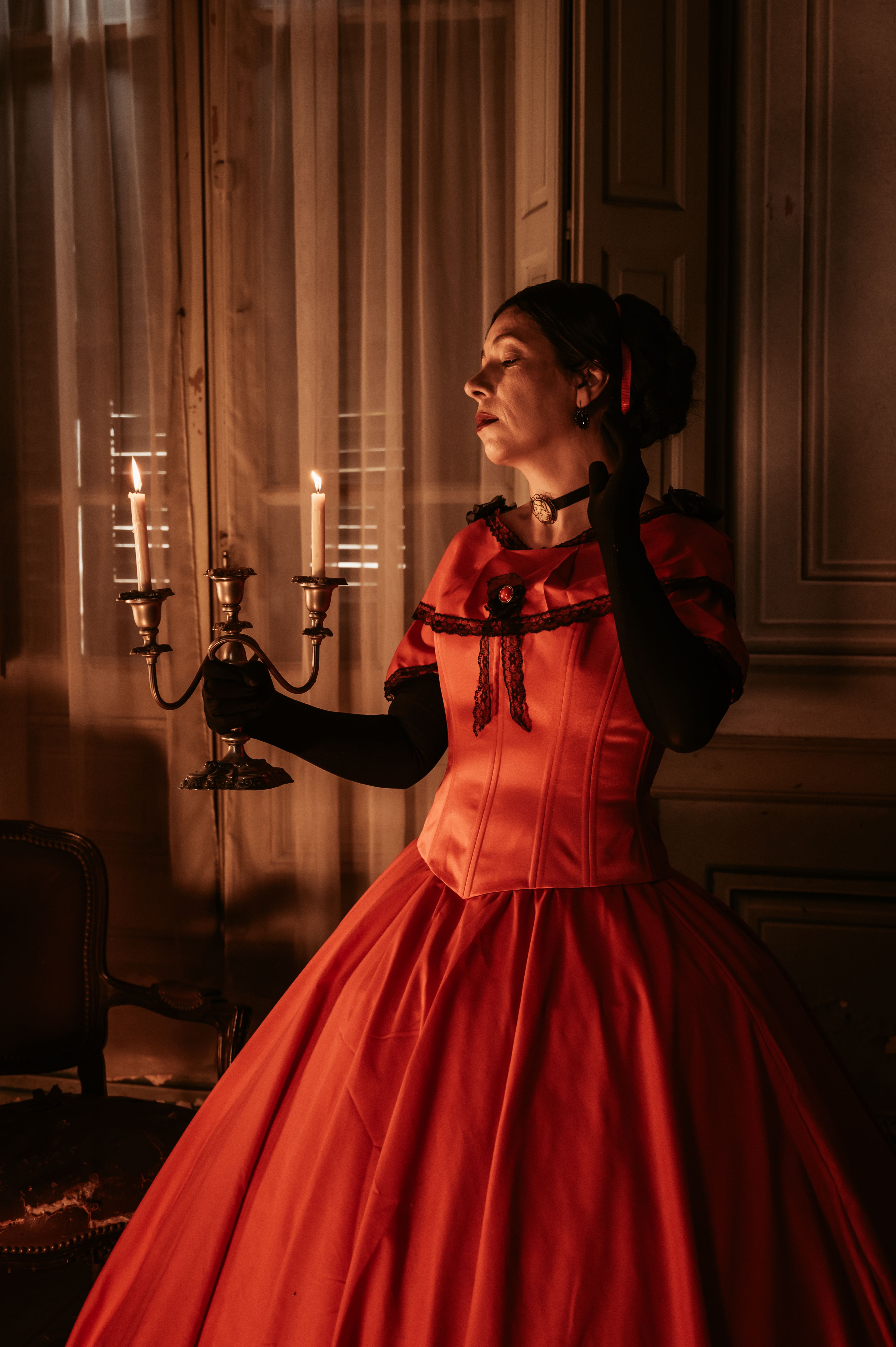 A woman in a vintage red gown holds a candlestick in a dimly lit room, creating an elegant atmosphere.