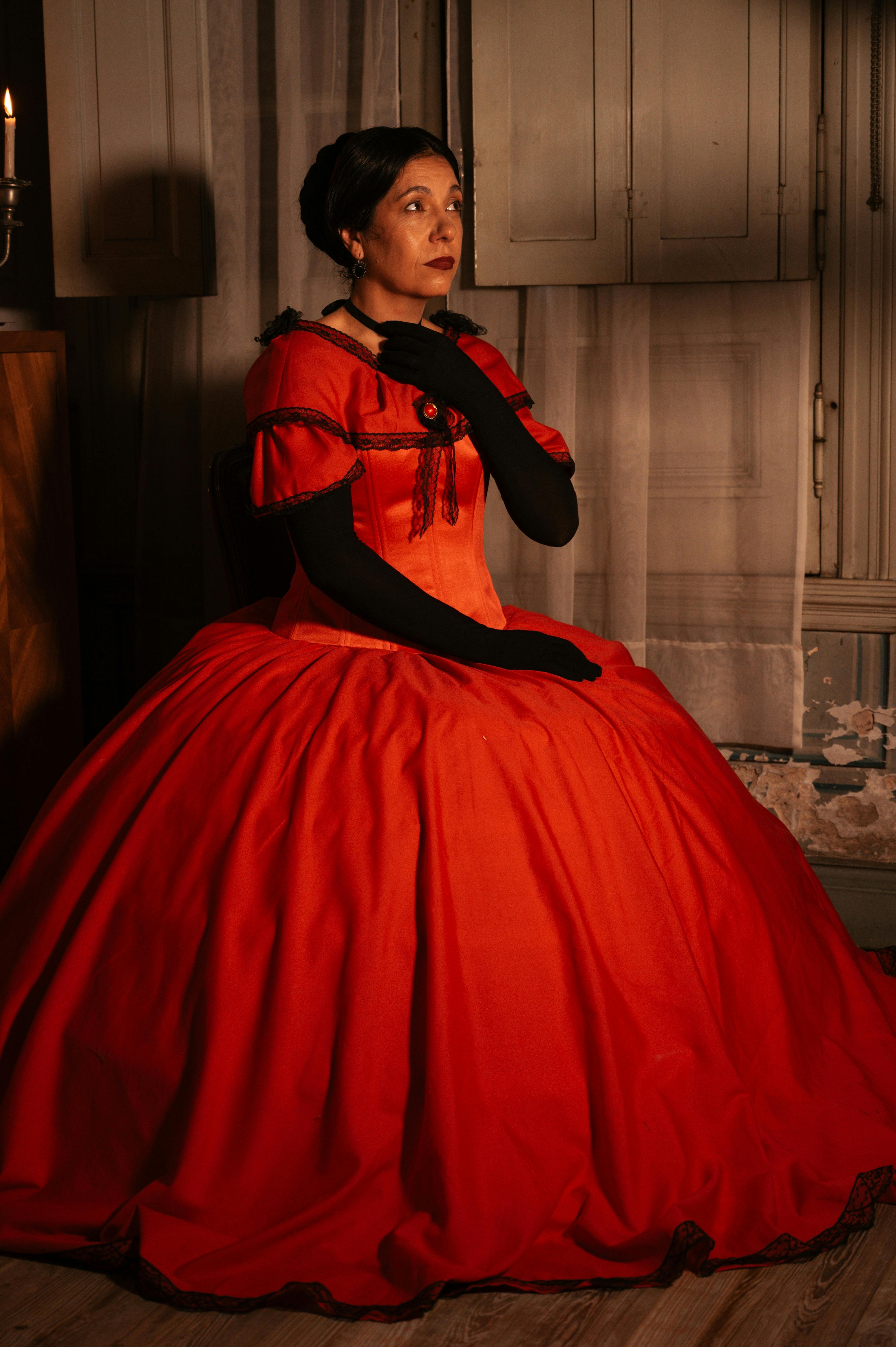 A woman wearing a historical red dress, seated indoors with soft lighting.