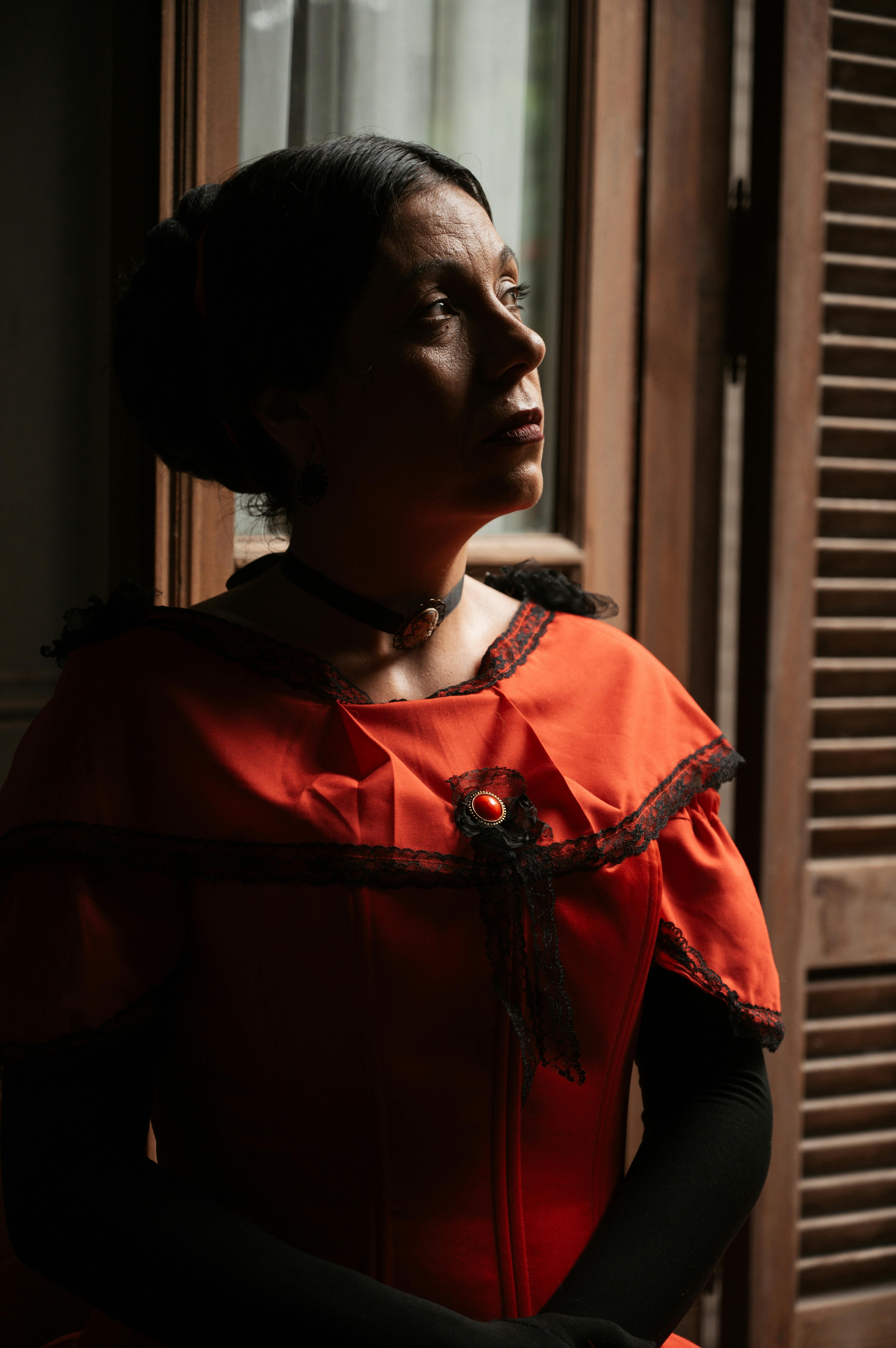 A woman in a vintage red dress looks thoughtfully out a window, accentuated by soft lighting.