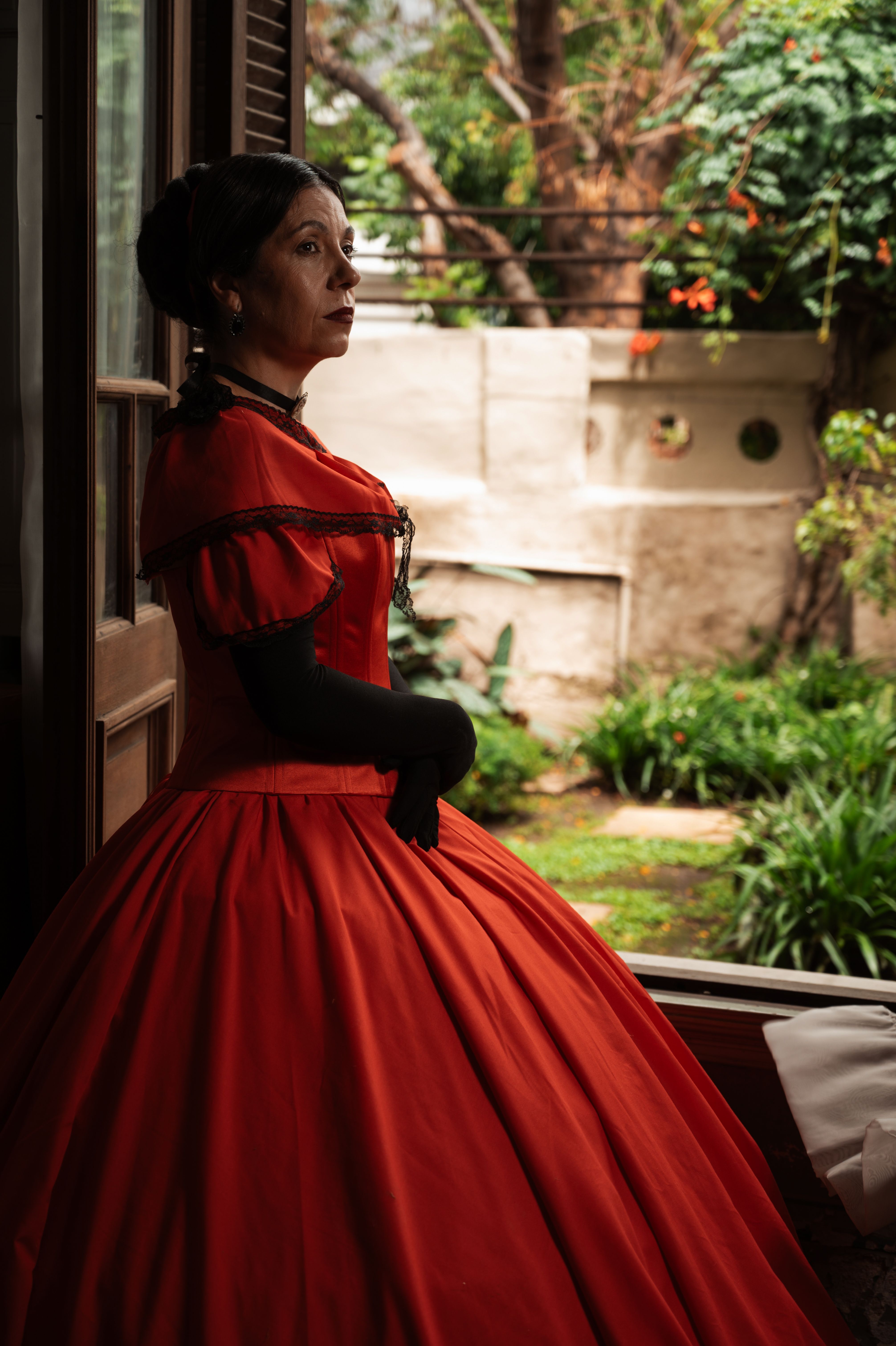 A woman in a red Victorian dress stands thoughtfully by a window, capturing a nostalgic ambiance.