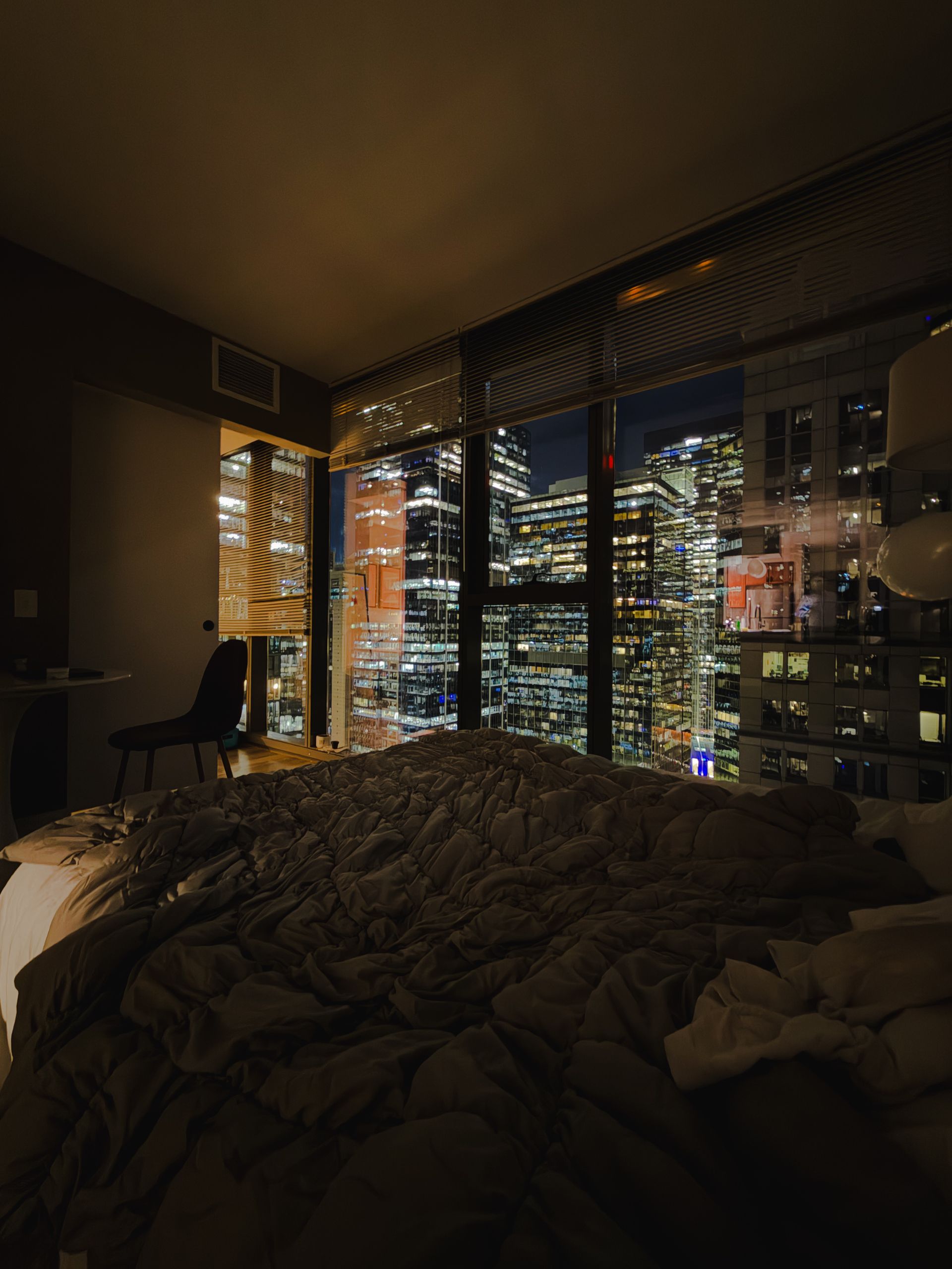 Dark bedroom at night, a rumpled bed facing tall windows full of lit city towers