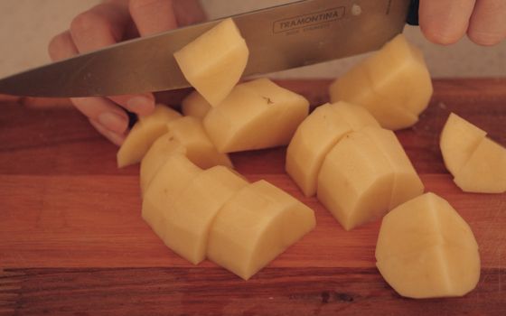 A person cuts potatoes on a wooden board with a kitchen knife.
