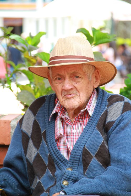 Free Portrait of an elderly man wearing an argyle sweater and hat, sitting outdoors with a thoughtful expression. Stock Photo