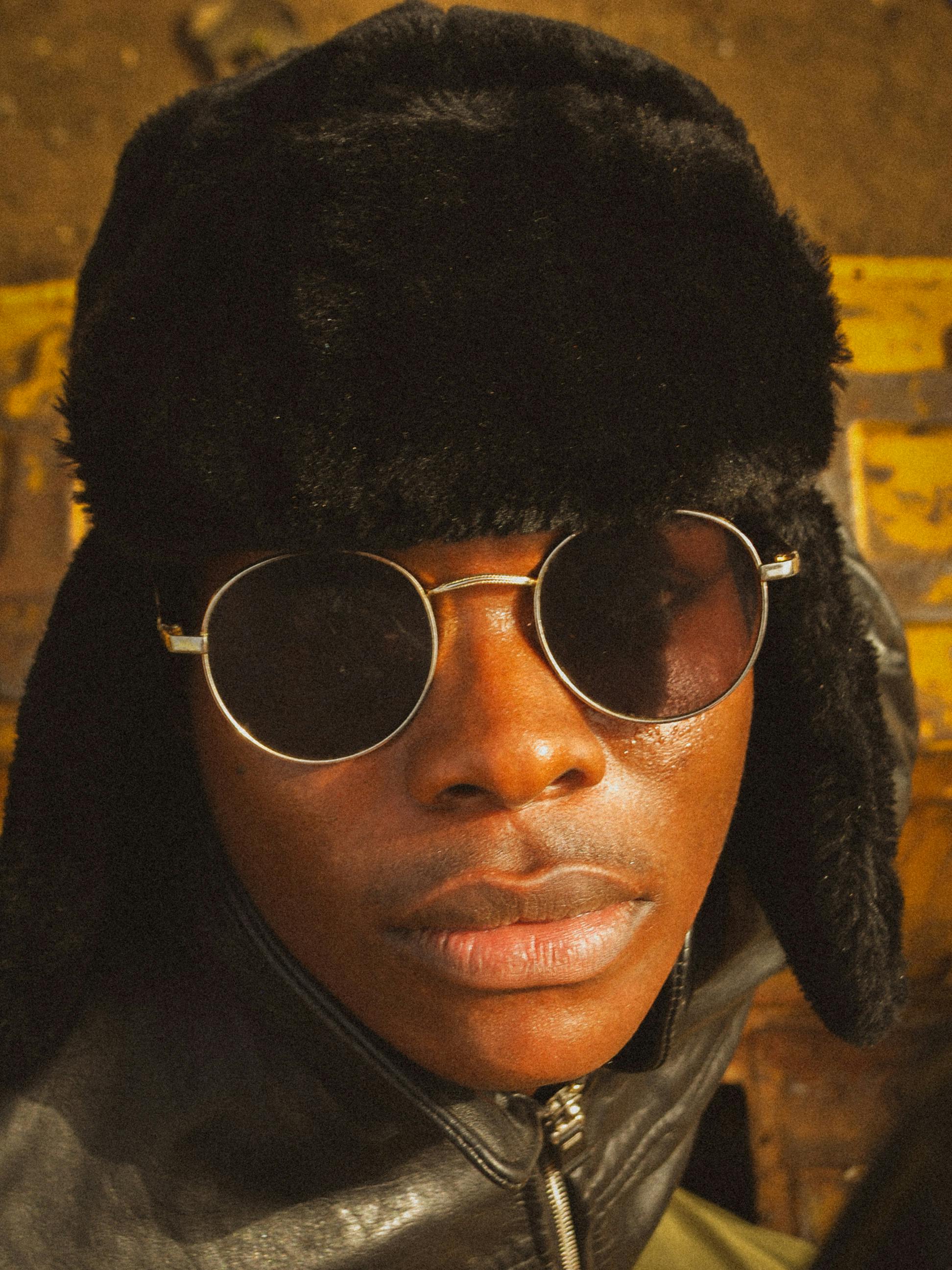 A young man wearing a fur hat and sunglasses in a close-up portrait, exuding a cool vibe.