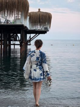 A woman in a floral dress walks barefoot along Alanya beach at sunset, holding sandals.