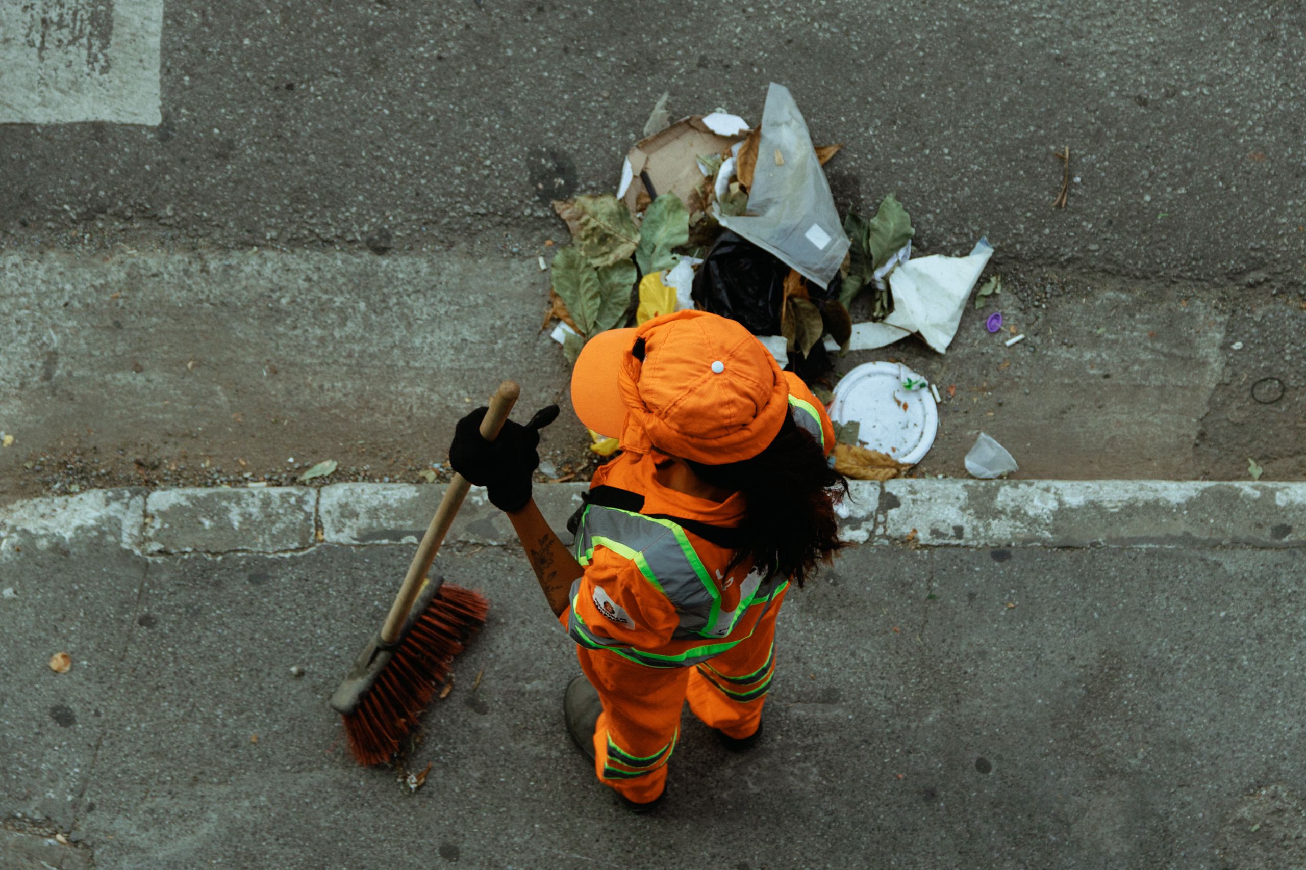 Top view of a street cleaner wearing orange uniform sweeping debris on a city sidewalk.
