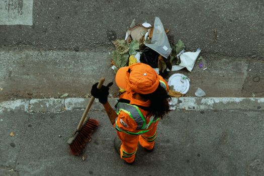 Top view of a street cleaner wearing orange uniform sweeping debris on a city sidewalk.
