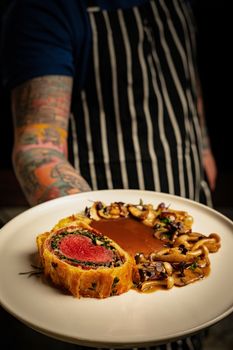 Delicious beef Wellington with mushrooms and sauce served by tattooed chef in apron.