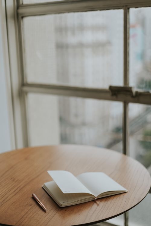 Free An open journal with a pen on a wooden table by a window, offering a cozy writing space. Stock Photo