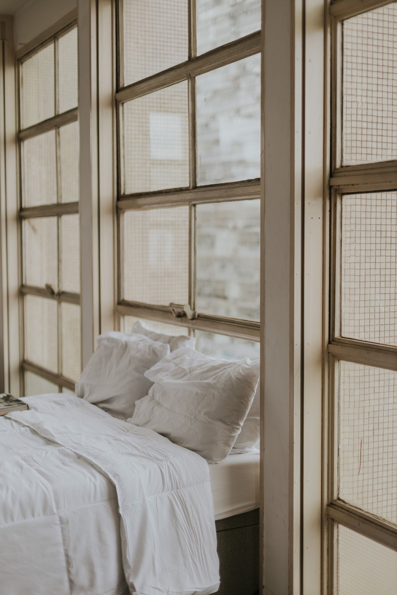Bed with crisp white bedding beside tall industrial windows in a small city apartment
