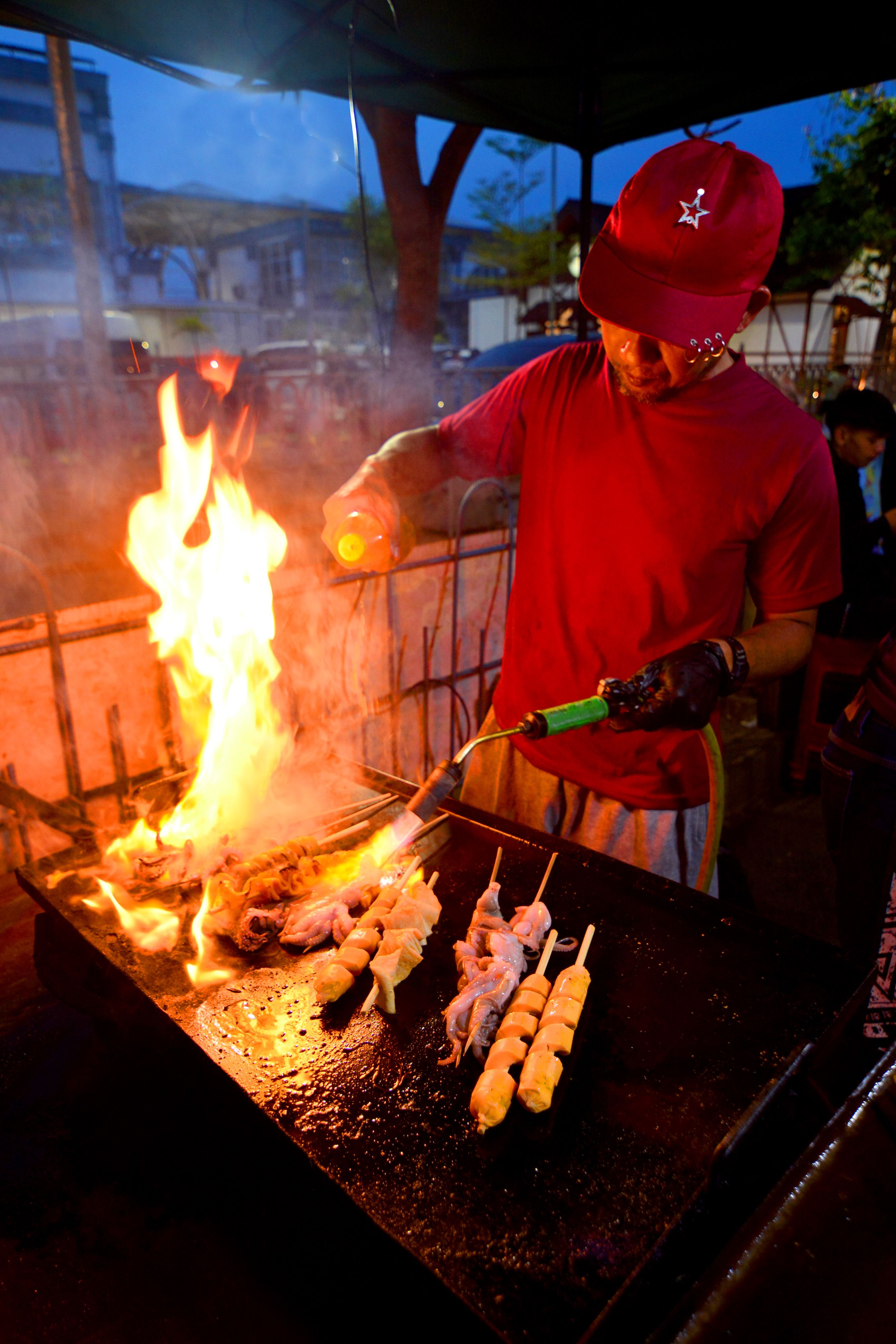 Dynamic scene of a street food vendor grilling skewers over open flames at a night market in West Java, Indonesia.
