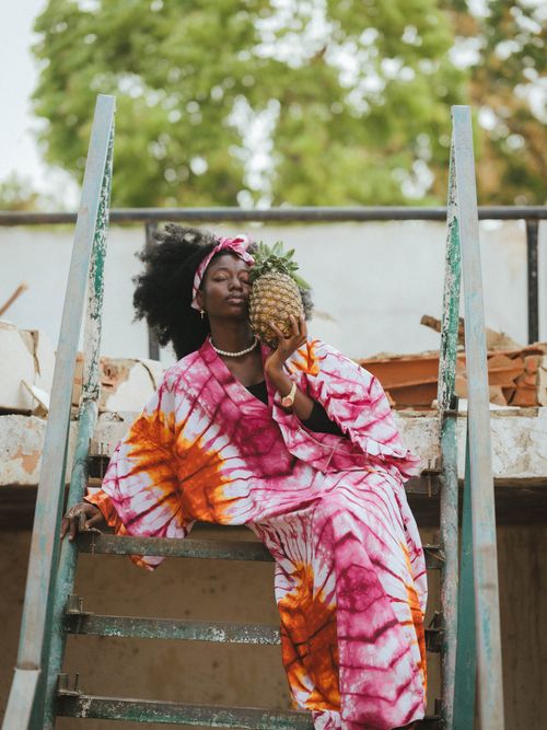 Free Young woman in colorful tie-dye dress holding a pineapple outdoors in Zaria City, Nigeria. Stock Photo