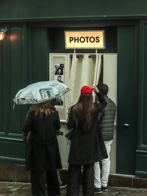 Three people wait outside a photobooth in Paris, holding an umbrella.