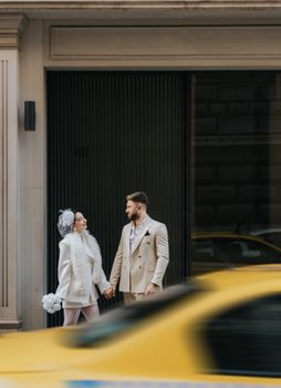 Elegant couple in fashionable attire holding hands in city with moving taxi.