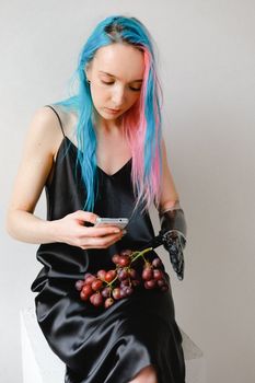 Young woman with colorful hair uses phone with prosthetic arm, grapes on lap.