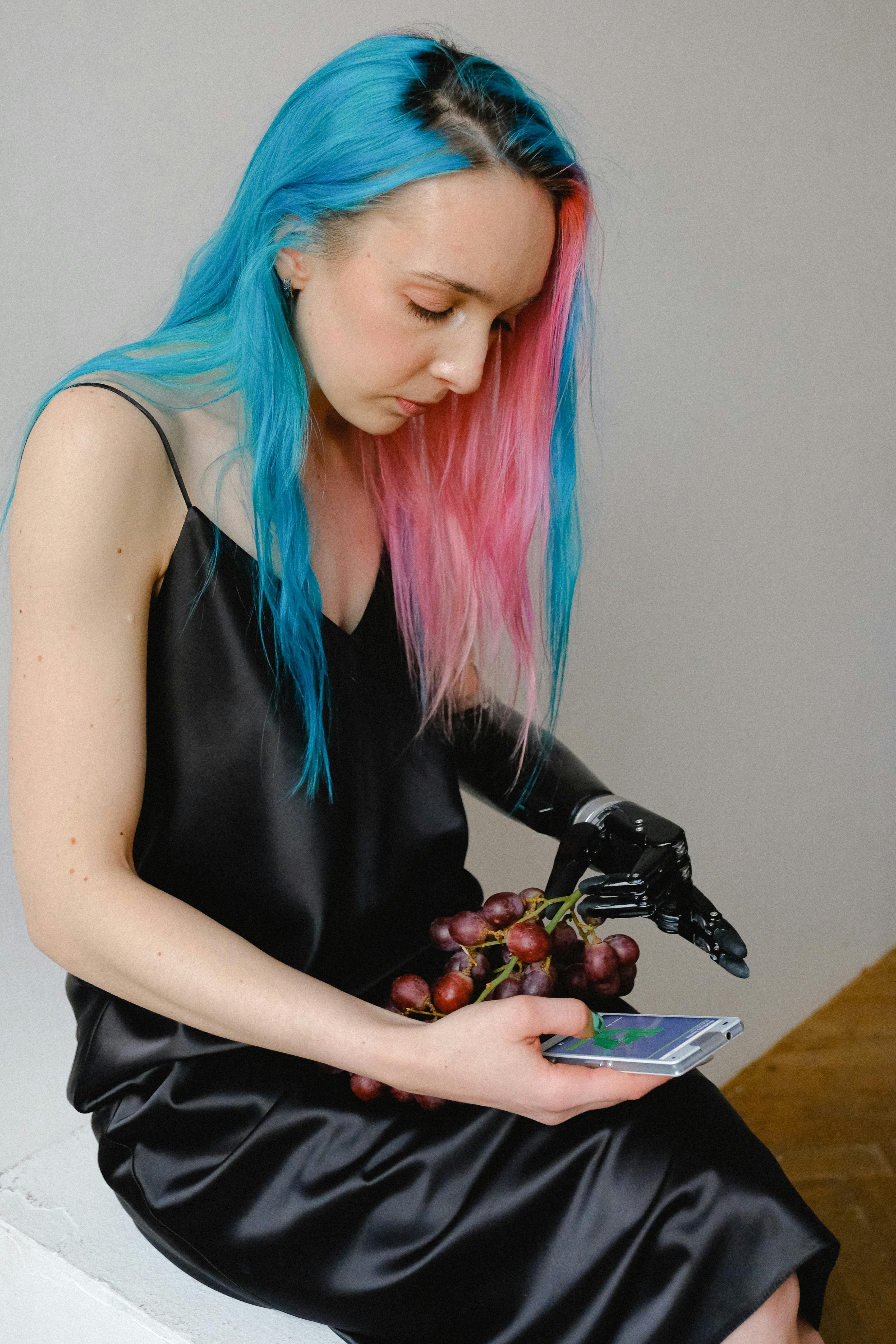 一位染着彩色头发的年轻女子一边使用智能手机，一边拿着葡萄，展现了包容性与现代生活方式