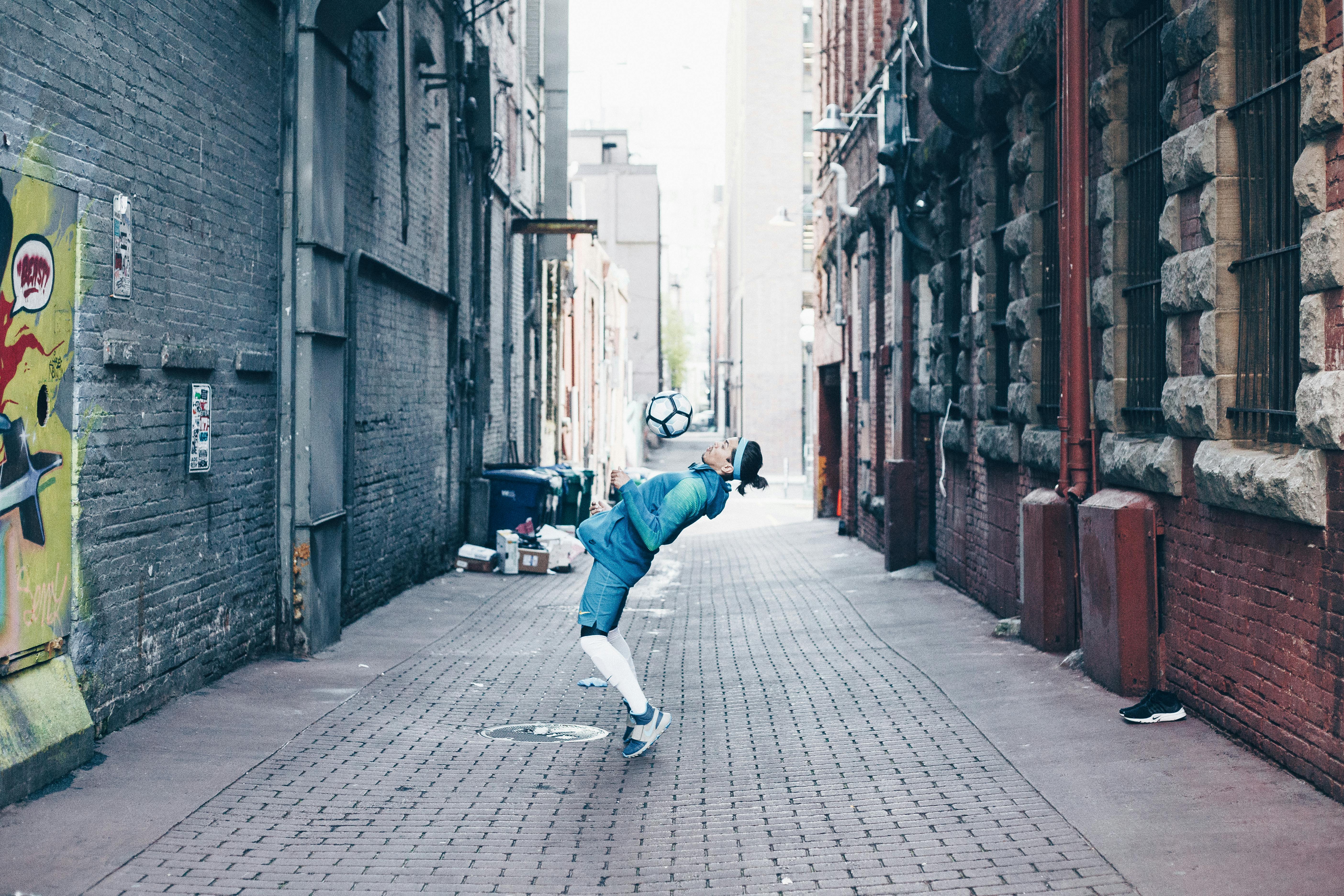 man in blue jacket and blue denim jeans playing football on an alley