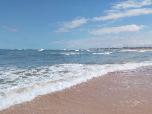 Free Waves gently lapping the shore on a serene sandy beach under a clear blue sky with distant city skyline. Stock Photo