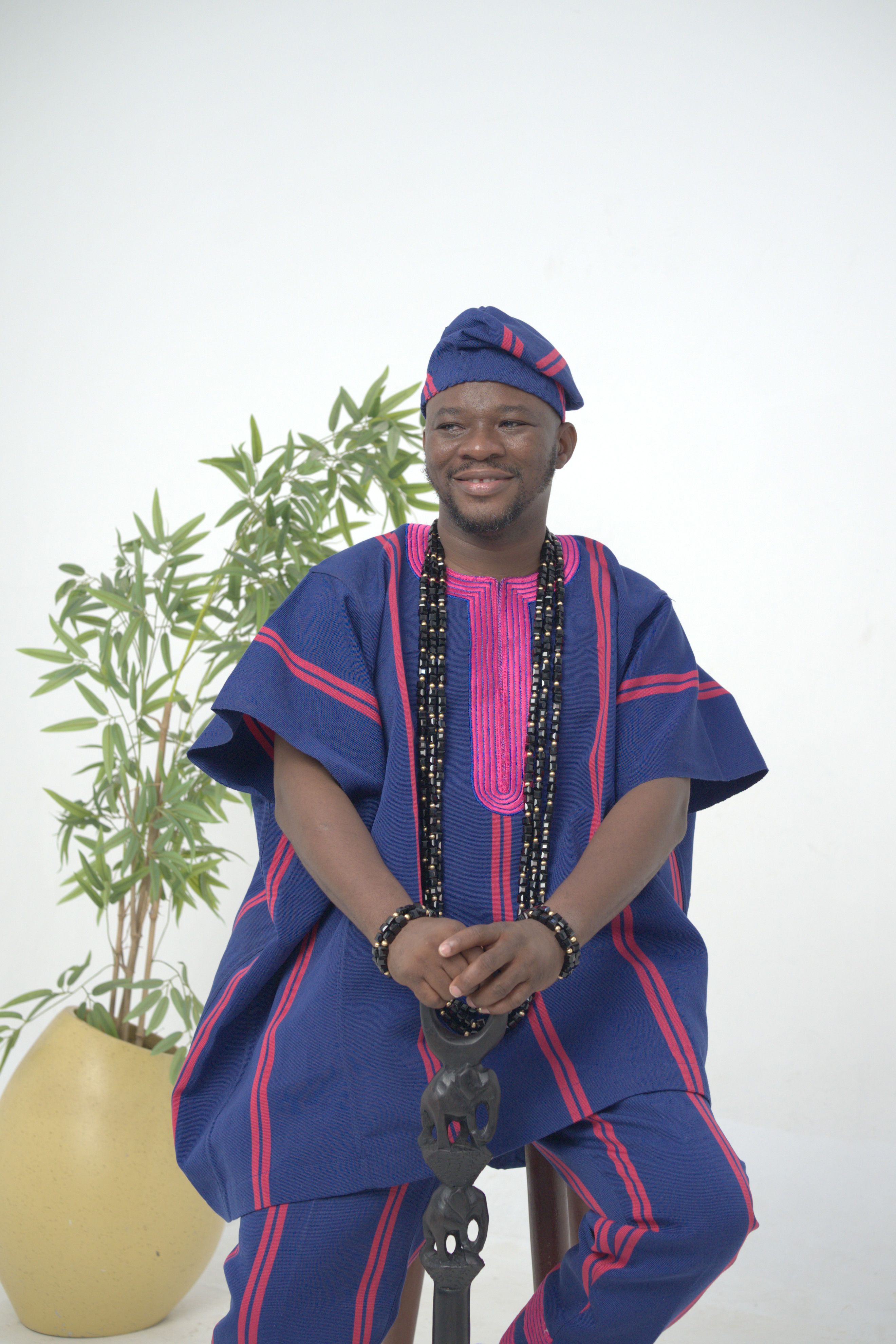 A smiling man in Nigerian traditional attire sits indoors, showcasing cultural fashion.