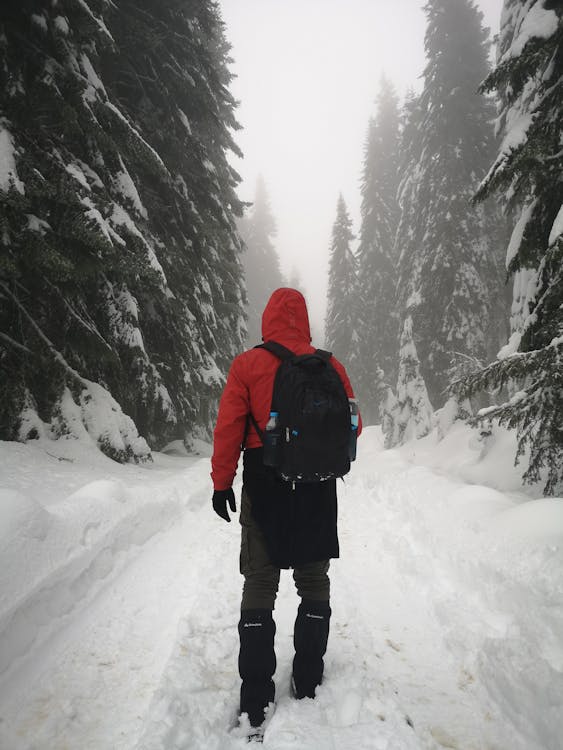 Person in Red Jacket and Black Pants Standing on Snow Covered Ground Carrying Backpack