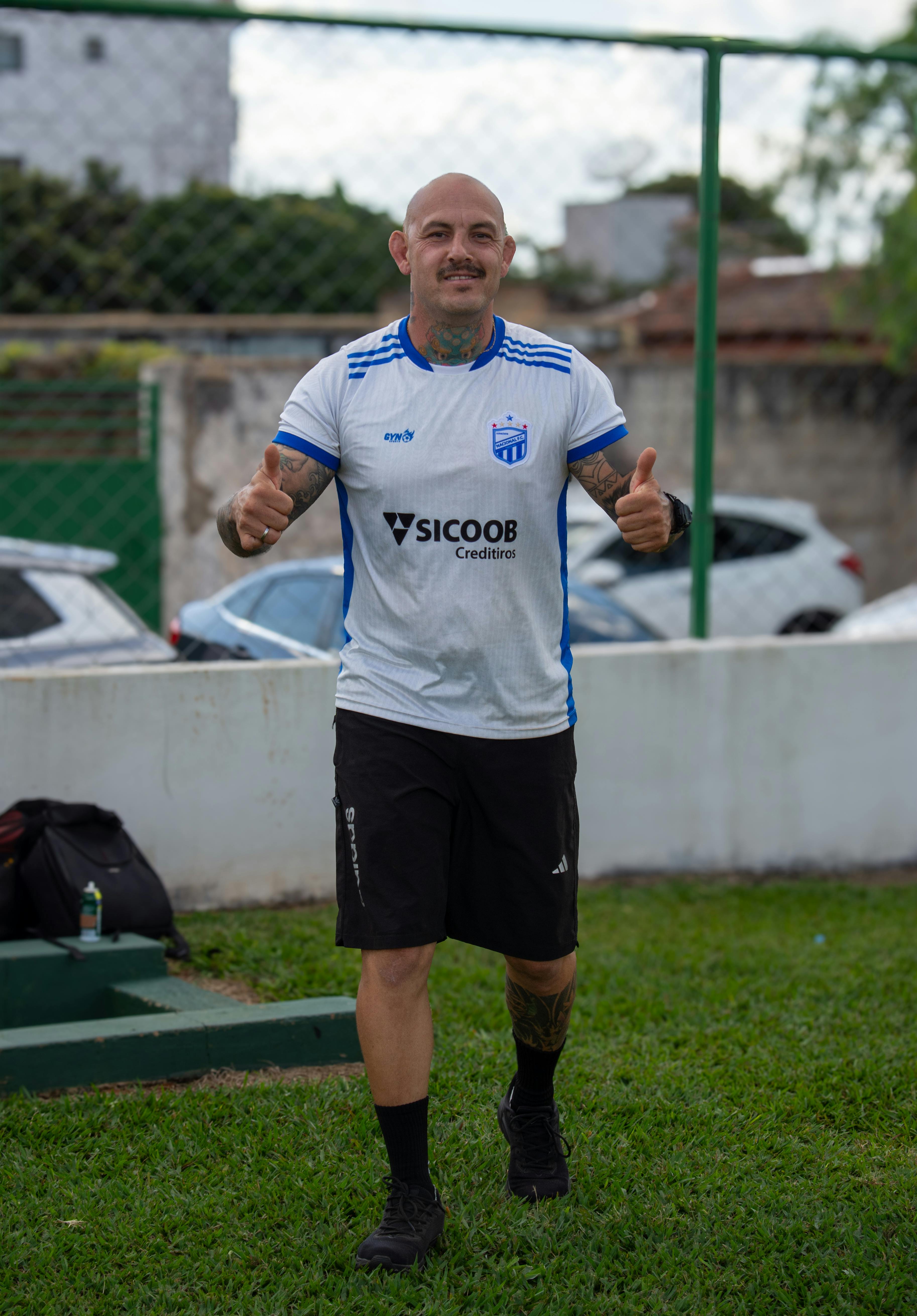 Smiling man in a soccer jersey giving thumbs-up outdoors on a sunny day.